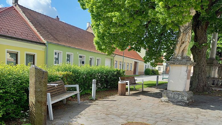 A quiet square with benches and a statue under a large tree, surrounded by colorful houses.