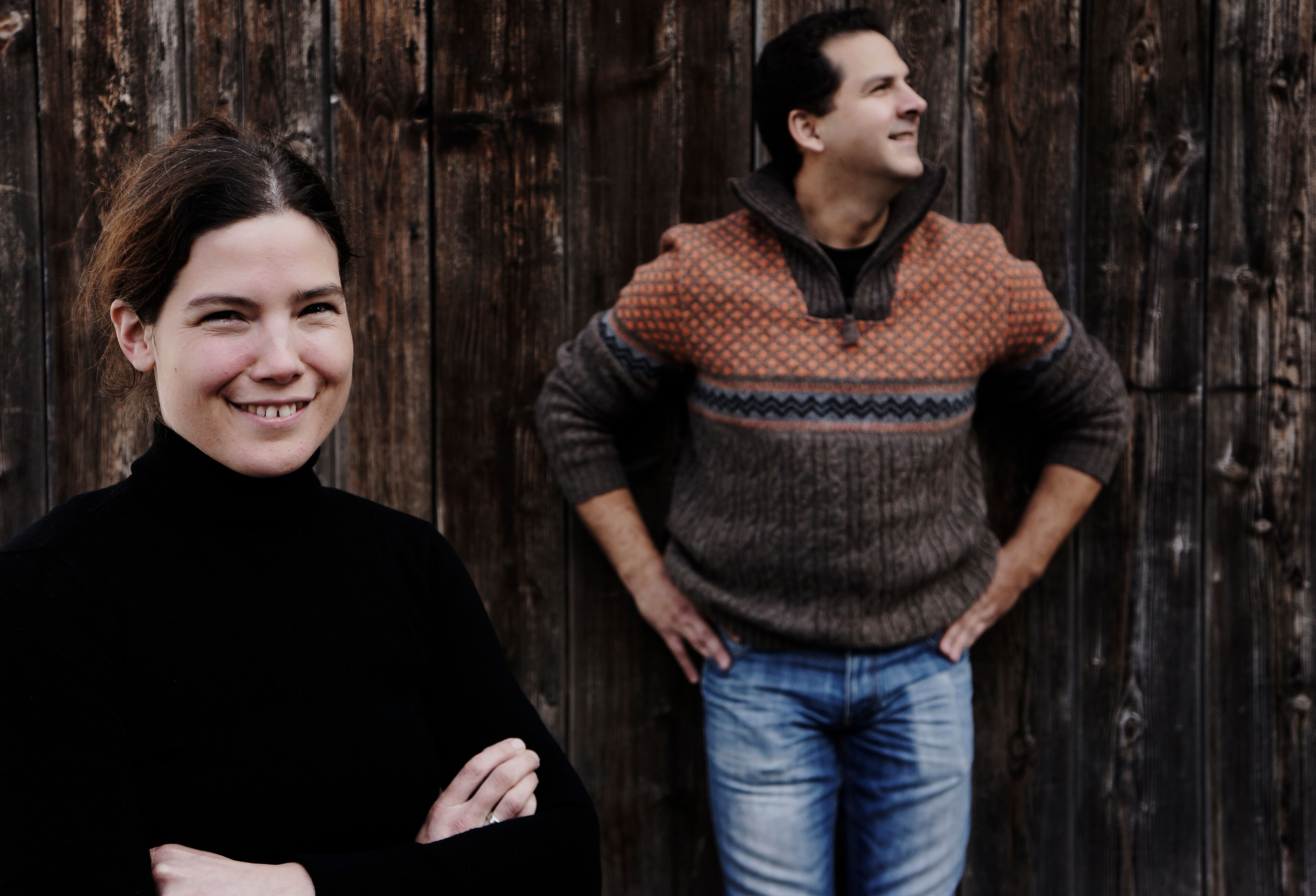 Zwei Personen vor einer Holzwand, eine Frau im Vordergrund lächelt, ein Mann im Hintergrund schaut zur Seite.