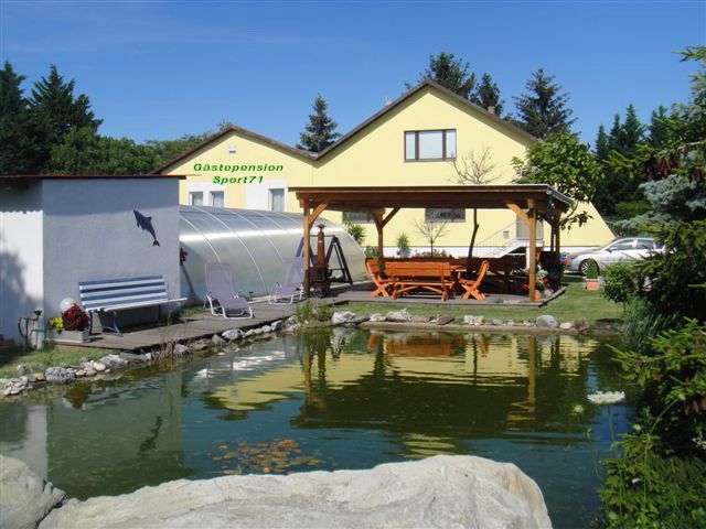 Exteriérový pohľad na žltý penzión s rybníkom a pavilónom.