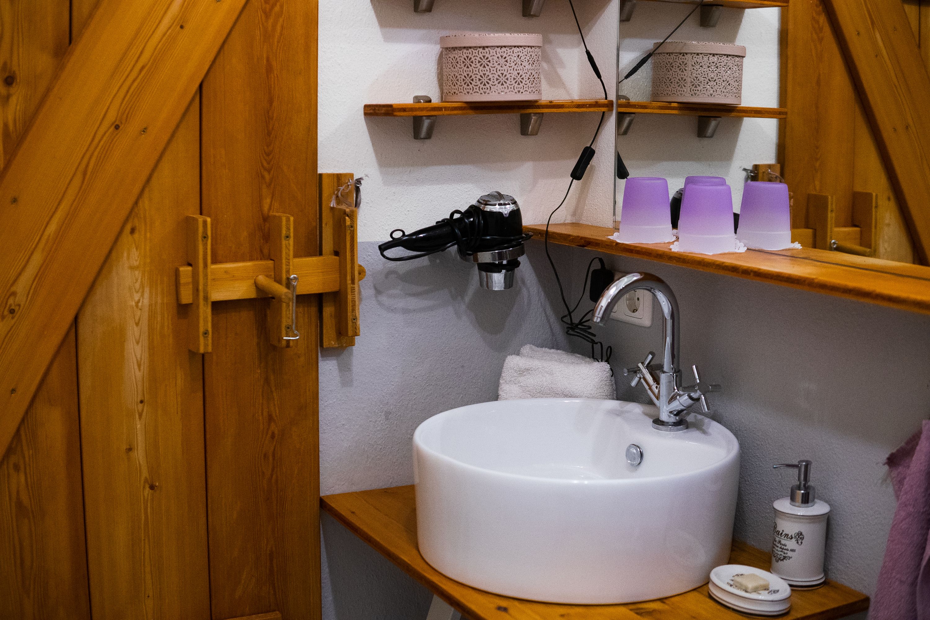 Badezimmer mit Waschbecken, Spiegel, Föhn und Holzregalen.