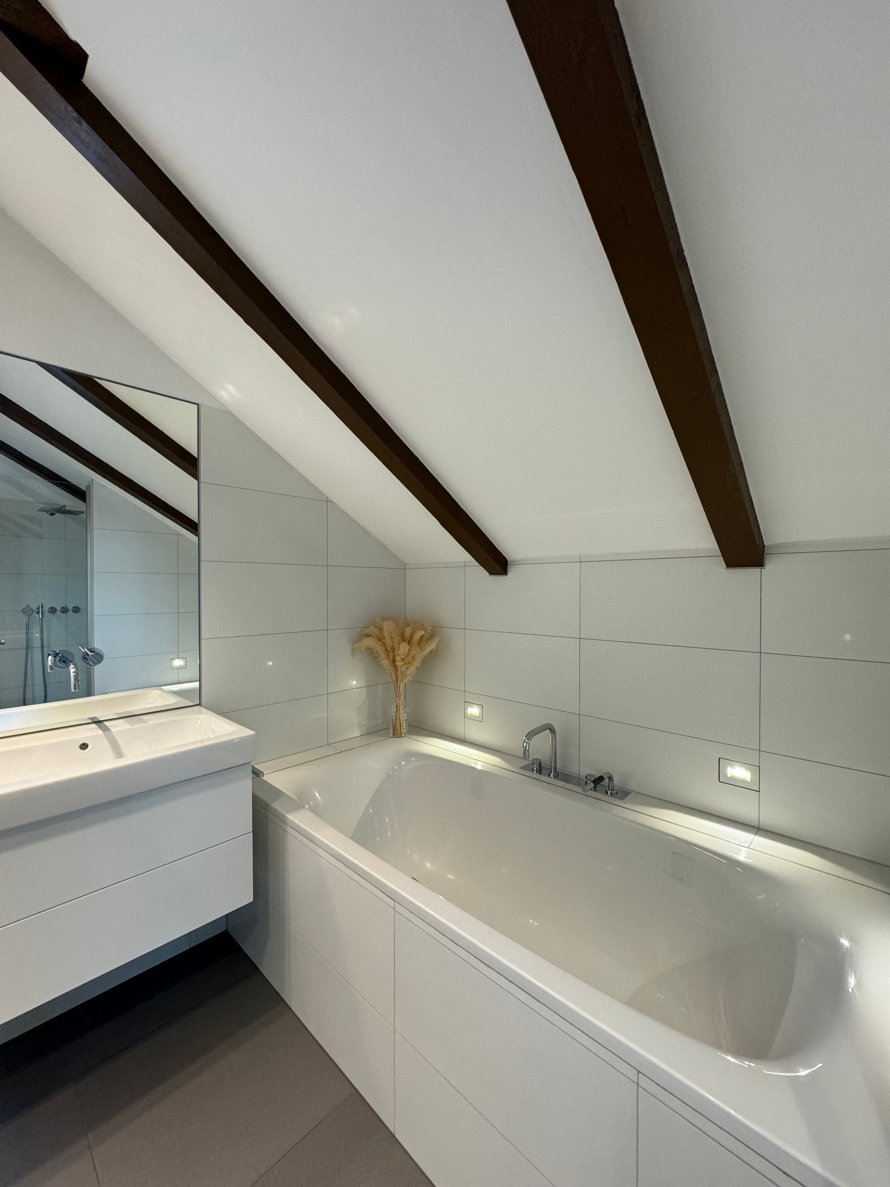 Modernes Badezimmer mit Badewanne, Waschbecken und schräger Decke mit Holzbalken.