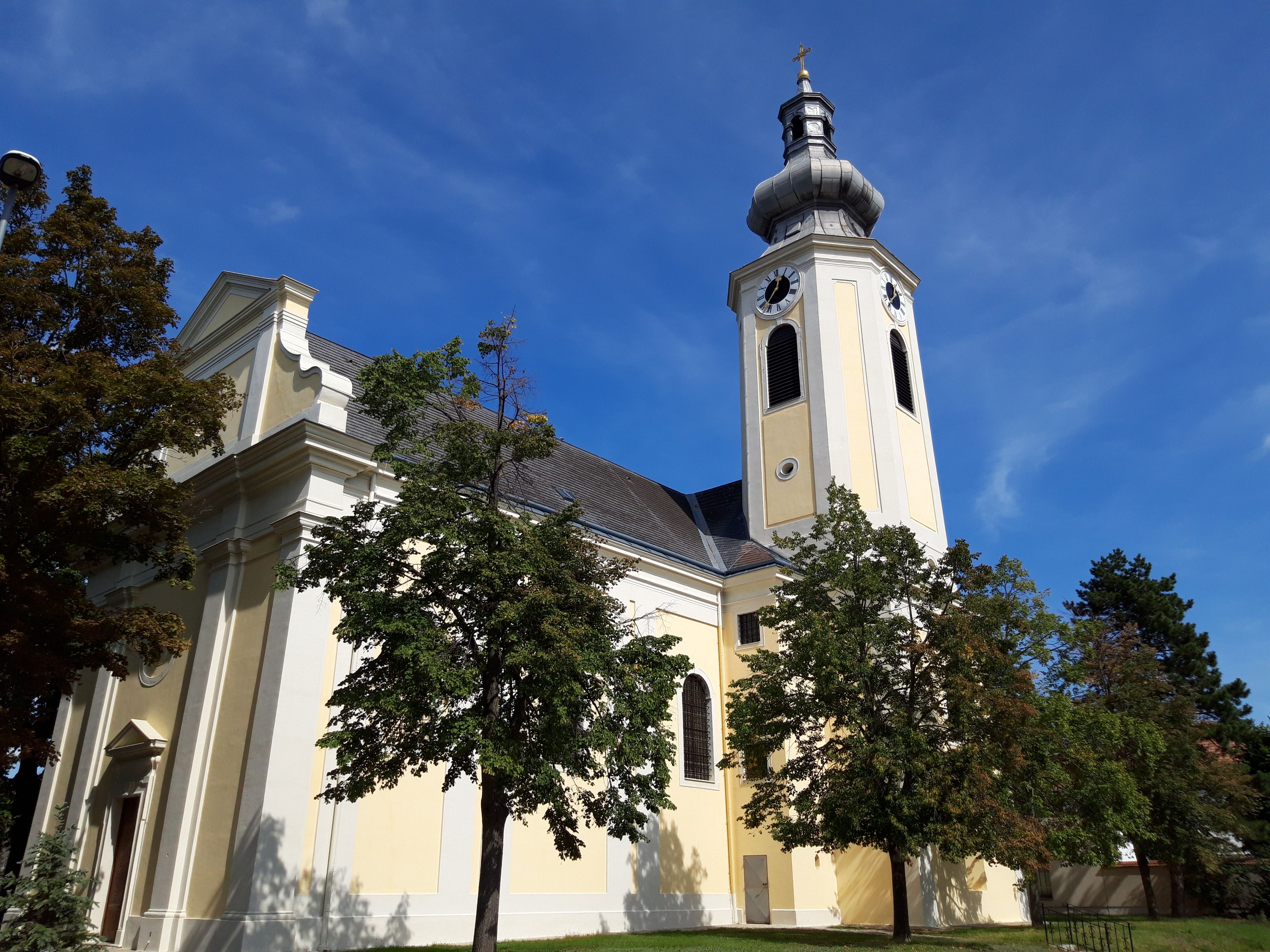 Gelbe Kirche mit Turm und Bäumen im Vordergrund unter blauem Himmel.