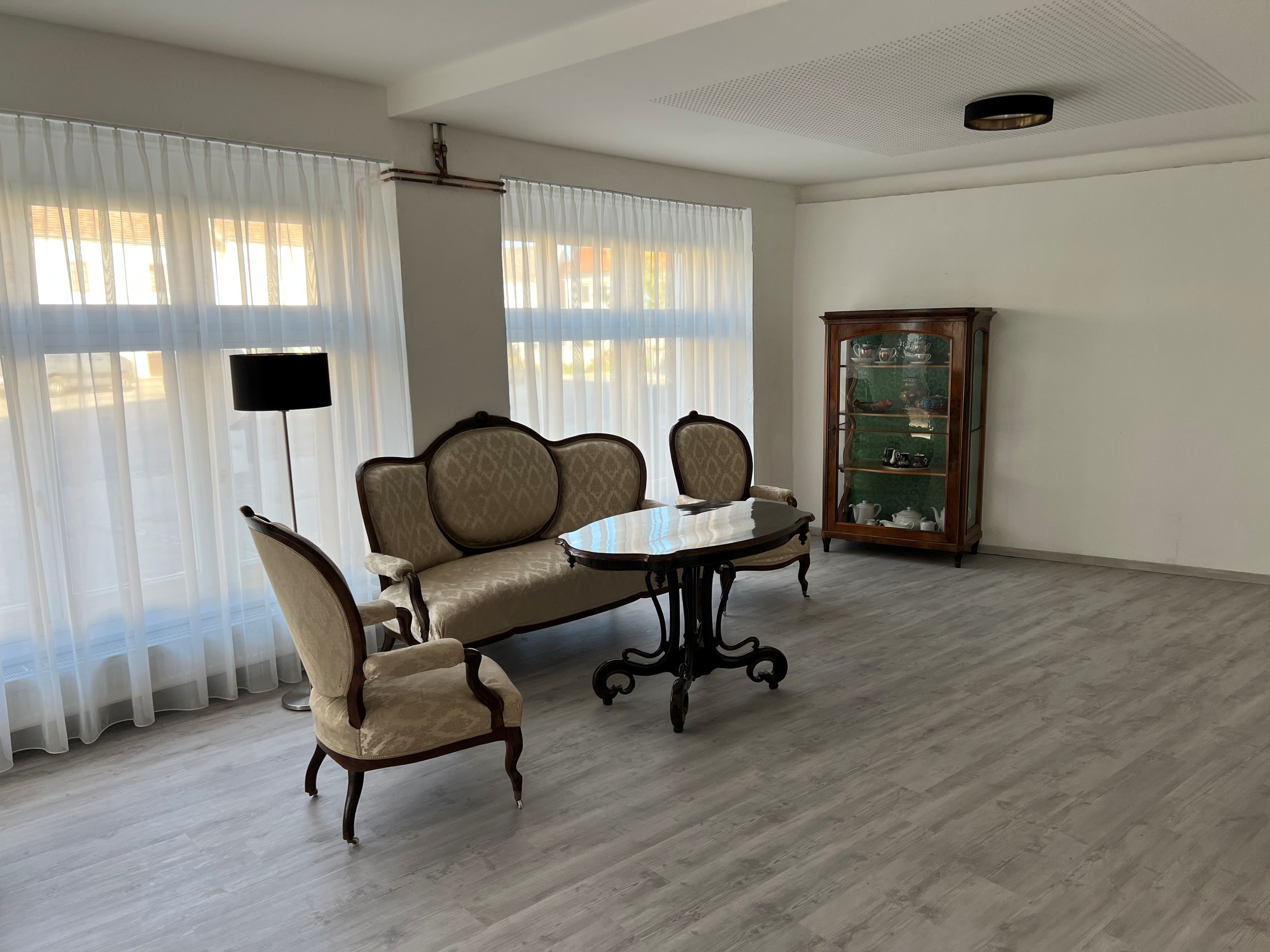 Ein eleganter Raum mit antiken Möbeln, darunter ein Sofa, ein Tisch und ein Schrank mit Geschirr. Große Fenster mit weißen Vorhängen lassen viel Licht herein.
