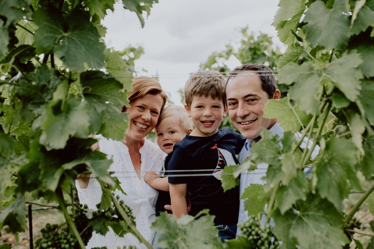 A smiling family stands among grapevines in a vineyard.