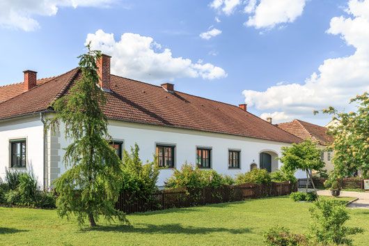 Weißes Haus mit rotem Ziegeldach, Garten und Bäumen unter blauem Himmel mit Wolken.