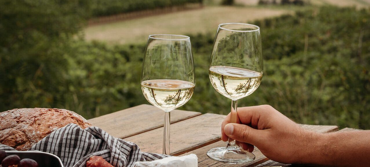 Auf einem Holztisch mit Blick auf die sanft-hügelige Weinviertler Landschaft ist eine Jause mit Hartwurst, Käse, Brot und Gemüse angerichtet. Eine Hand greift nach einem der beiden eingeschenkten Weißweingläser.