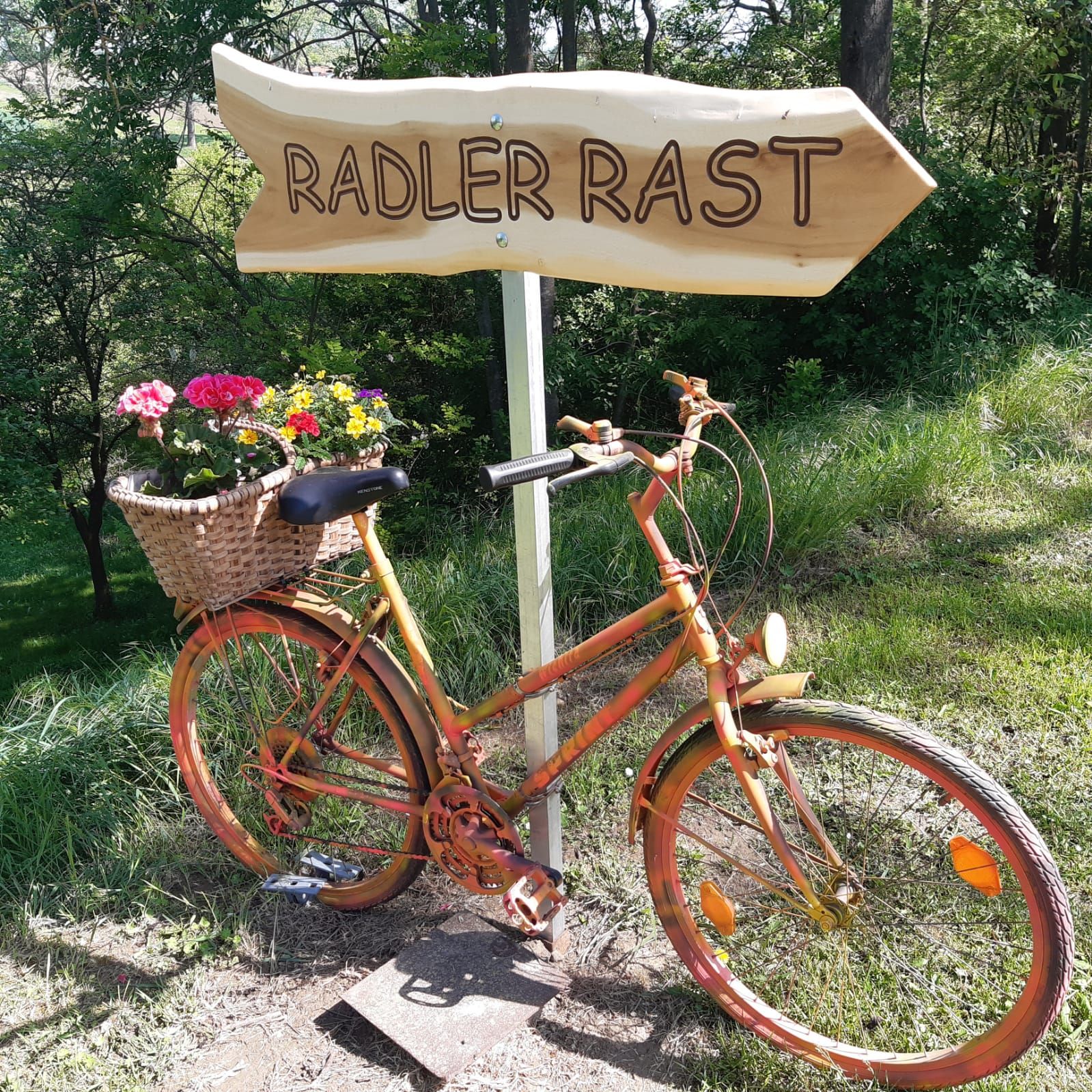 Ein orangefarbenes Fahrrad mit einem Blumenkorb steht neben einem Holzschild mit der Aufschrift 'Radler Rast' in einer grünen Umgebung.