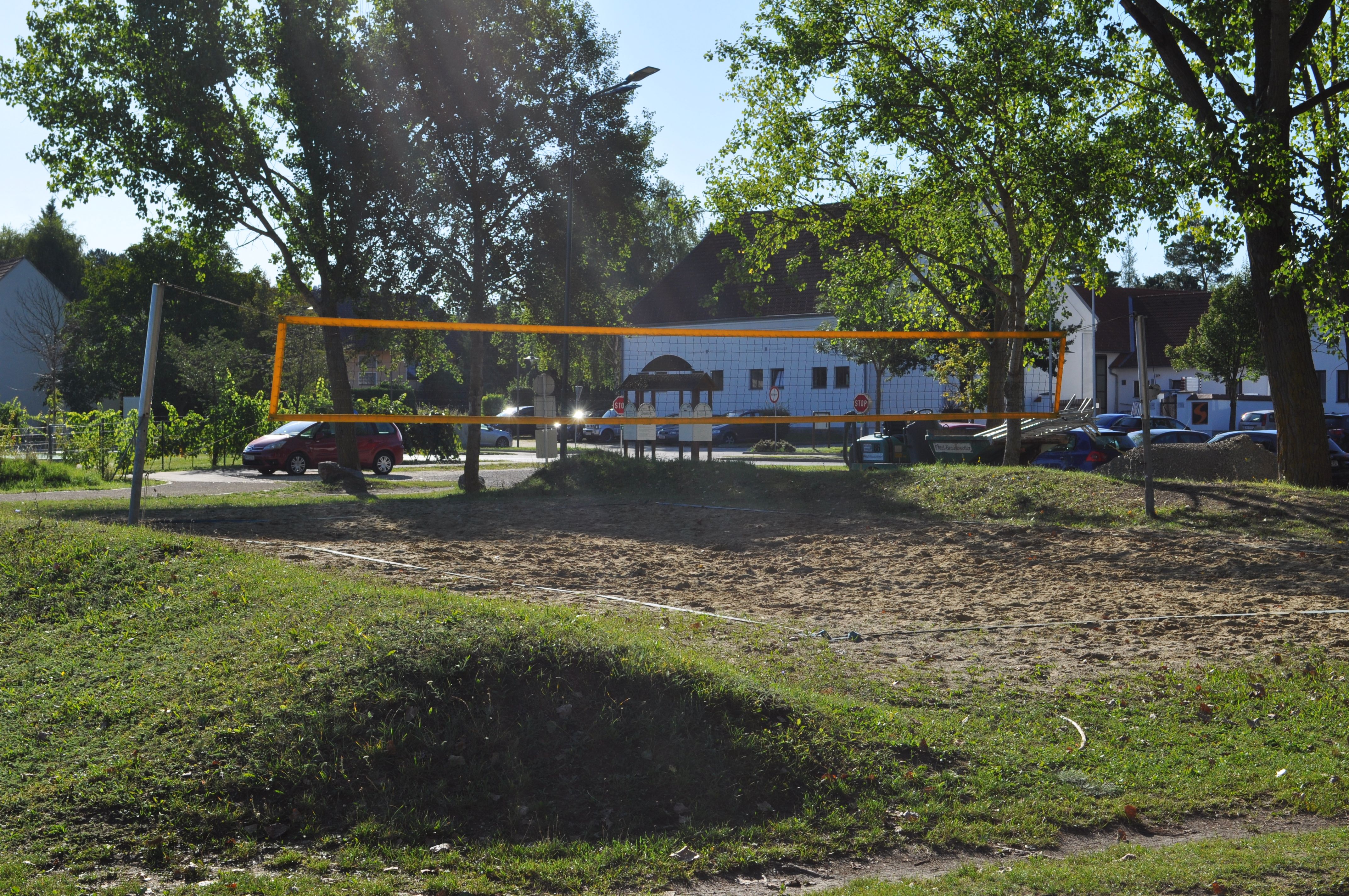 Ein leerer Volleyballplatz mit Netz, umgeben von Bäumen und parkenden Autos im Hintergrund.