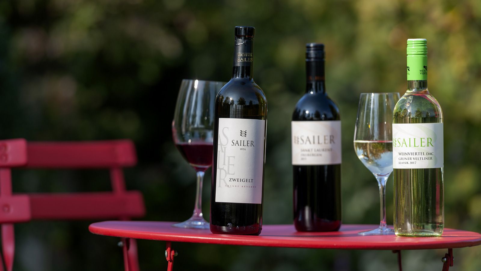 Drei Weinflaschen und zwei Weingläser auf einem roten Tisch im Freien.