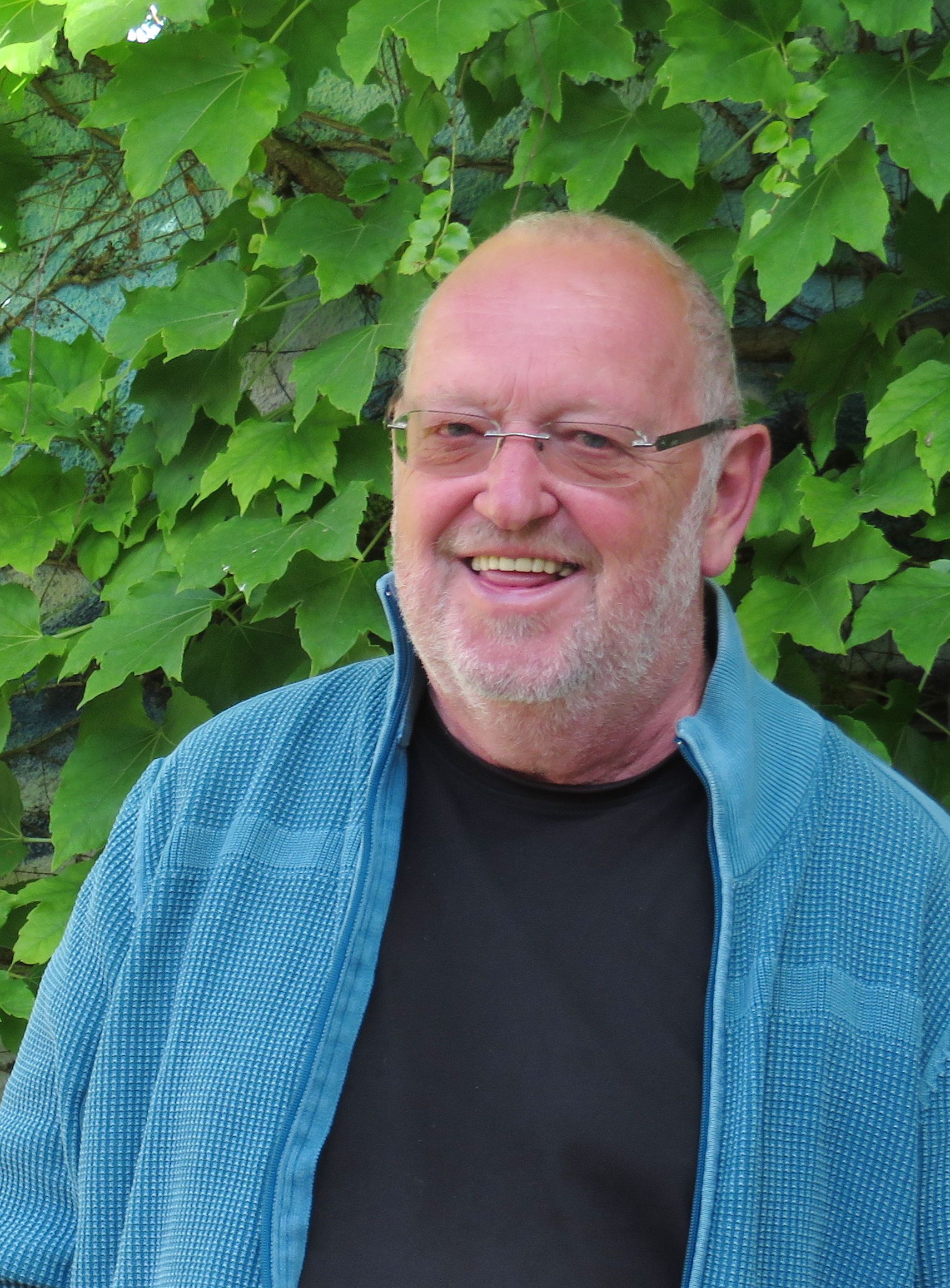 Ein älterer Mann mit Brille und Bart lächelt vor einer grünen Blätterwand. Er trägt eine blaue Jacke und ein schwarzes T-Shirt.