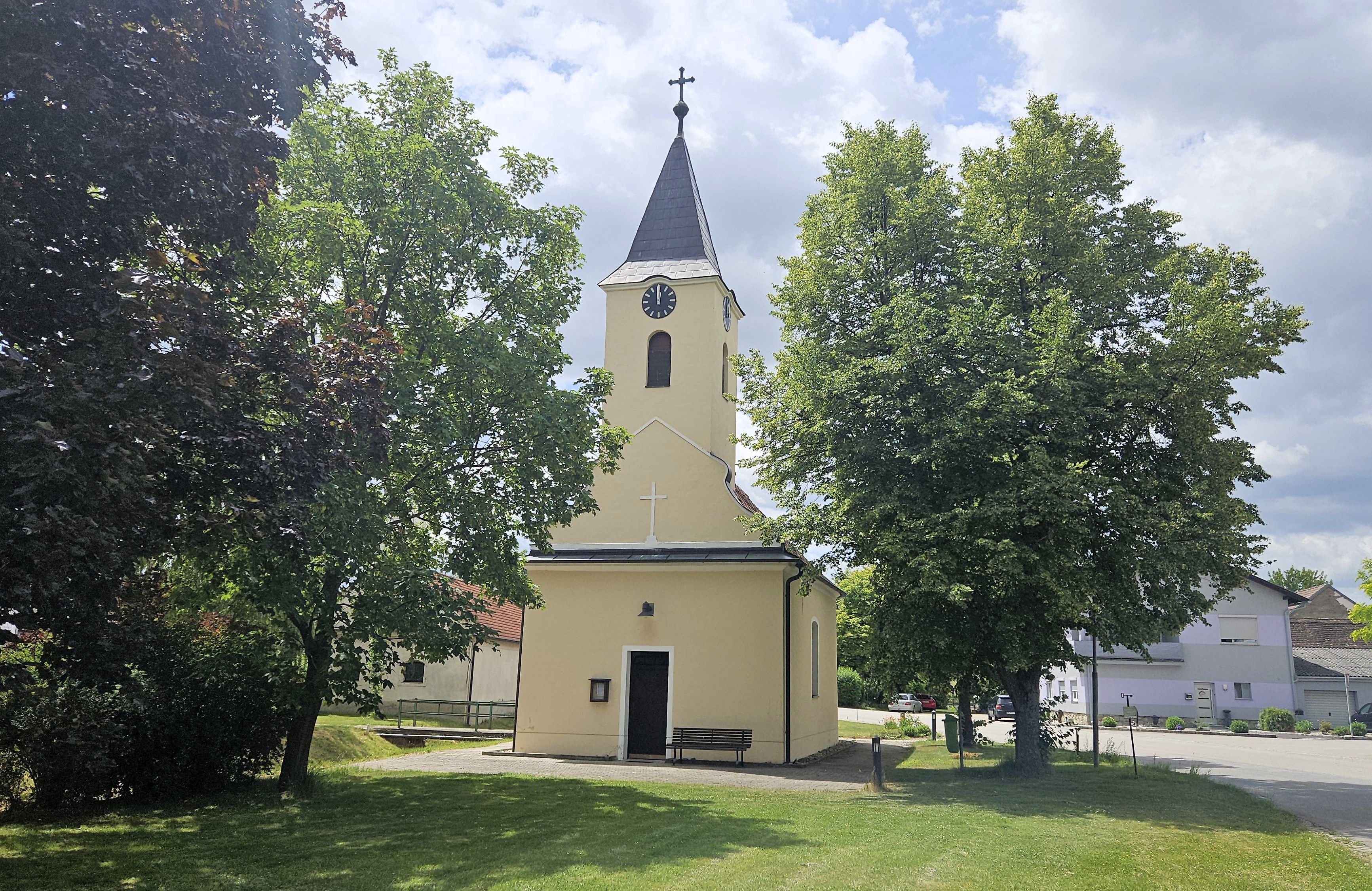 Die Barbara Kapelle in Kleinweikersdorf, umgeben von Bäumen und einem gepflegten Rasen, bei sonnigem Wetter.