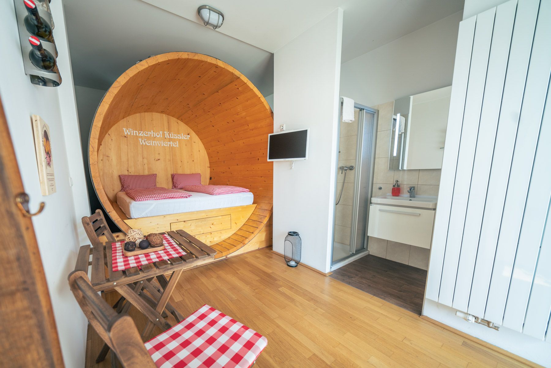 Zimmer im Winzerhof Küssler mit rundem Holzbett, kleinem Tisch mit rot-weiß karierten Decken und angrenzendem Bad.