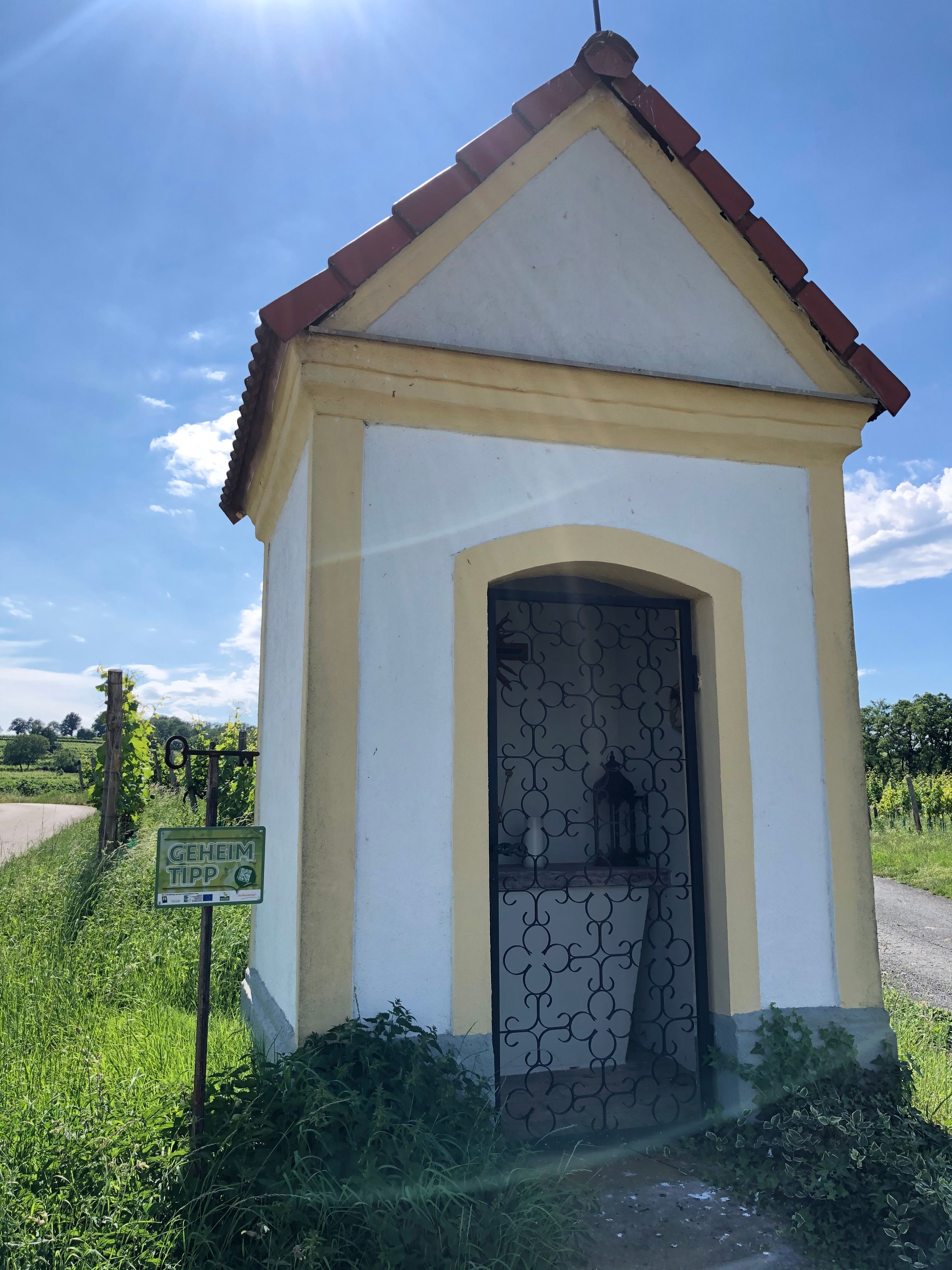 Weinbergkapelle von Glaubendorf mit schmiedeeisernem Tor und Schild 'Geheimtipp' daneben, umgeben von grüner Landschaft.