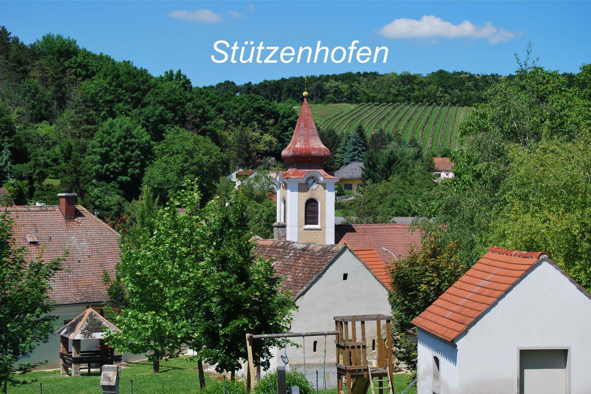 View of the village of Stützenhofen with a church and surrounding houses, surrounded by green trees and vineyards.