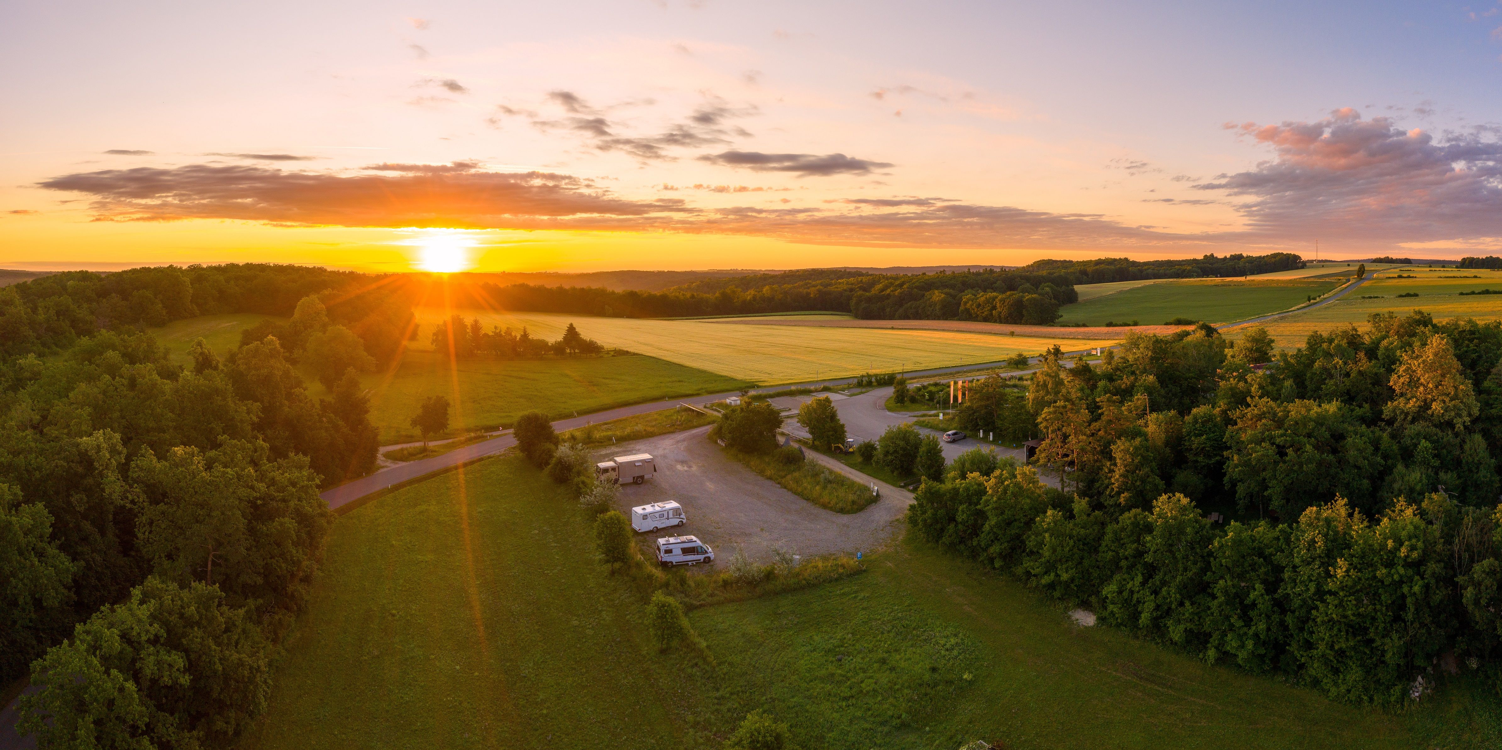 Luftaufnahme eines Wohnmobilstellplatzes bei Sonnenuntergang, umgeben von Feldern und Wäldern.