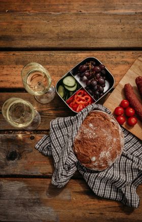 Auf einem Holztisch ist eine Jause mit zwei Gläsern Weißwein, einem Laib Brot, einer Box mit Trauben und Gemüse sowie einem Holzbrett mit Weinviertler Hartwurst angerichtet.