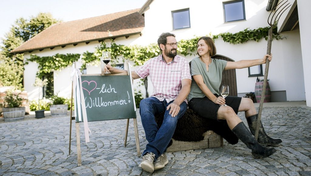 Ein Mann und eine Frau sitzen vor einem Haus mit einem Willkommensschild und Weingläsern.