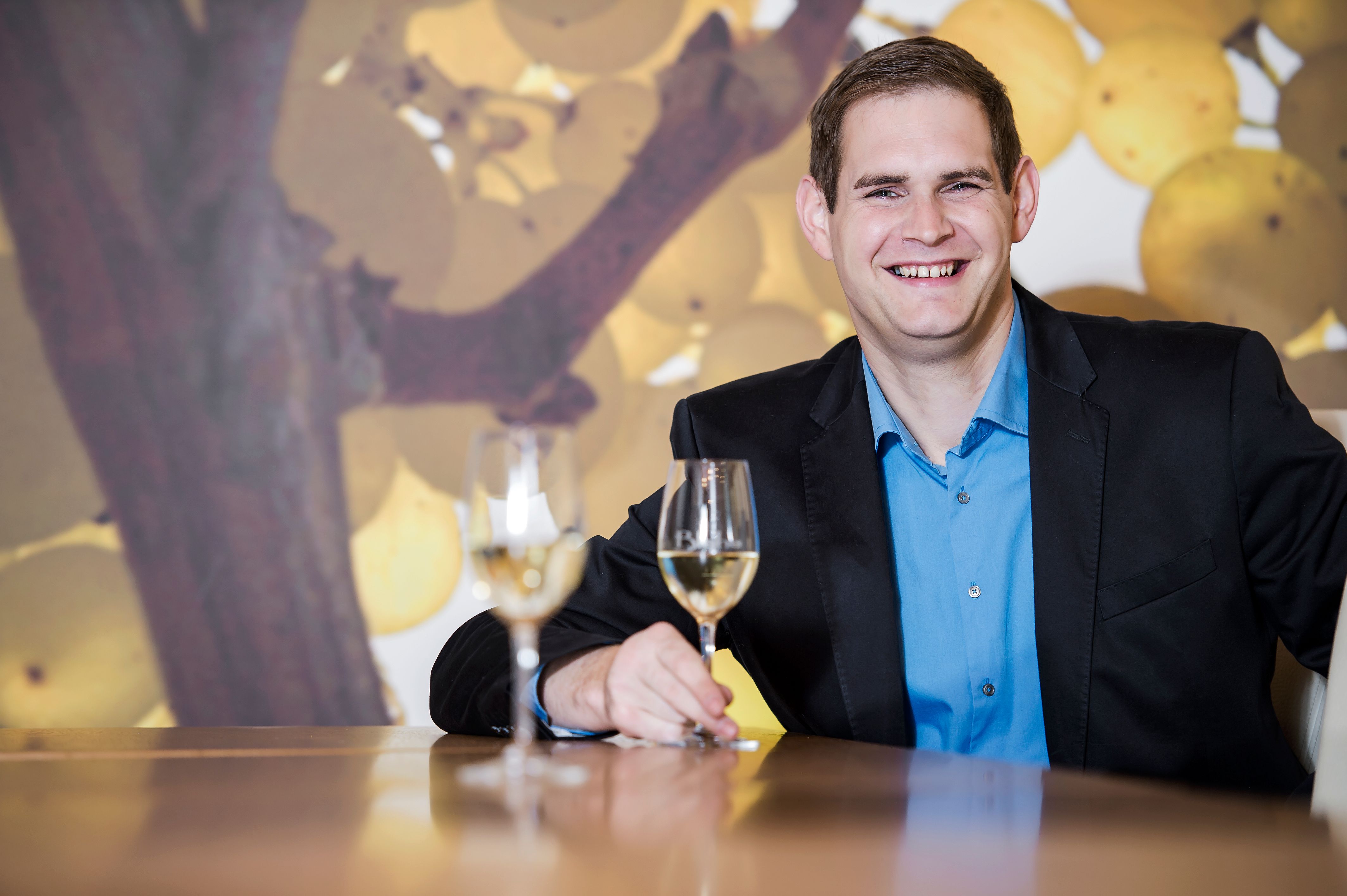 Ein Mann in einem Anzug sitzt an einem Tisch mit zwei Weingläsern vor einem Hintergrund mit Weintrauben.