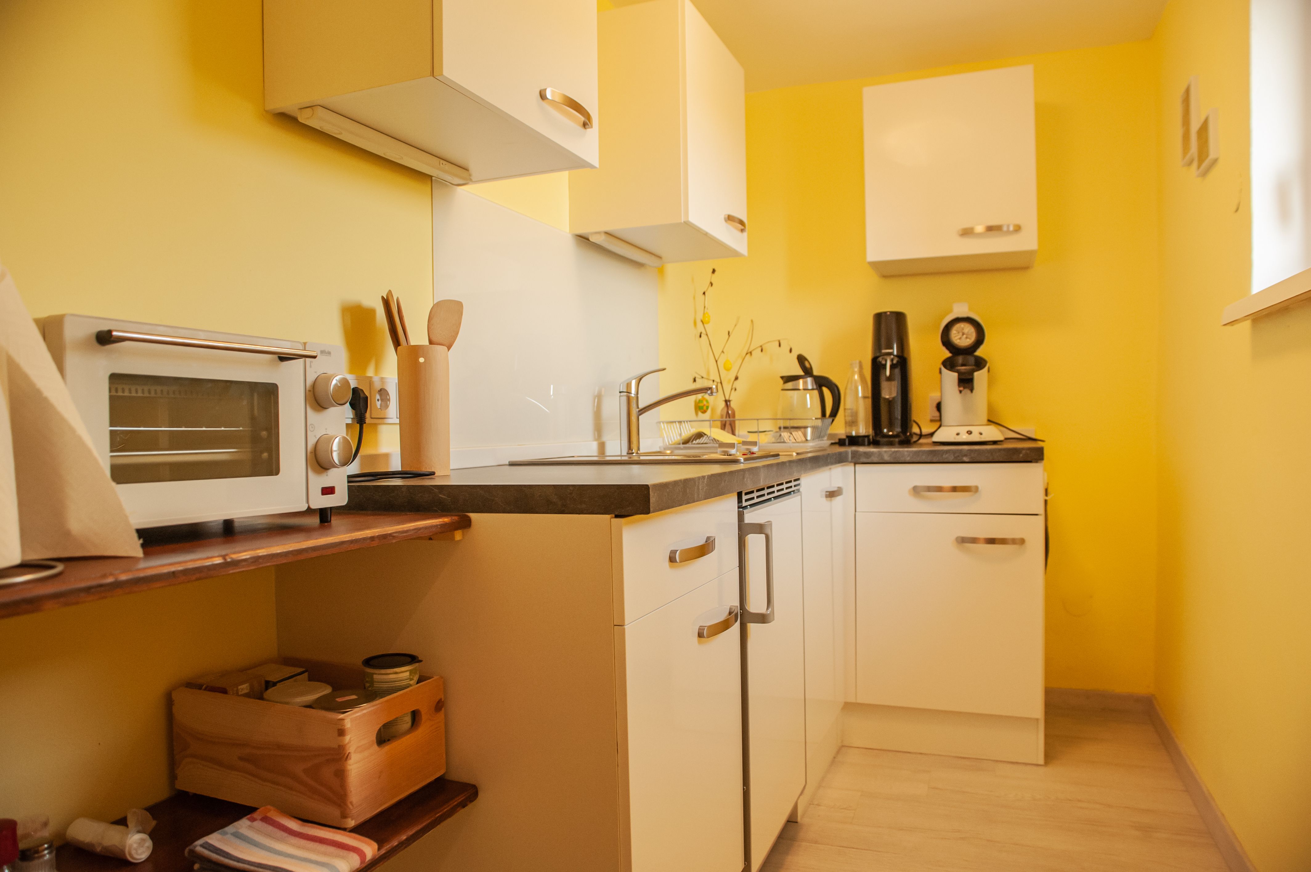 Malá kuchyňa so žltými stenami, bielymi skrinkami a spotrebičmi, ako je hriankovač a kávovar.