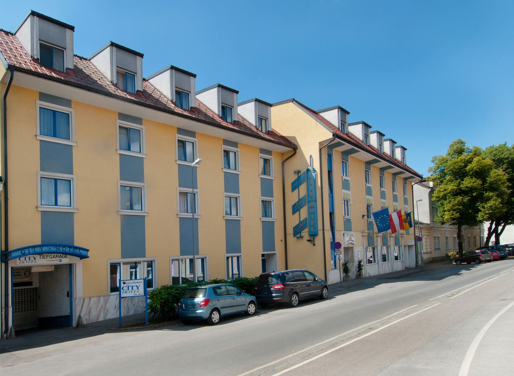 Außenansicht eines gelb-blauen Hotels mit mehreren Fenstern und geparkten Autos davor.