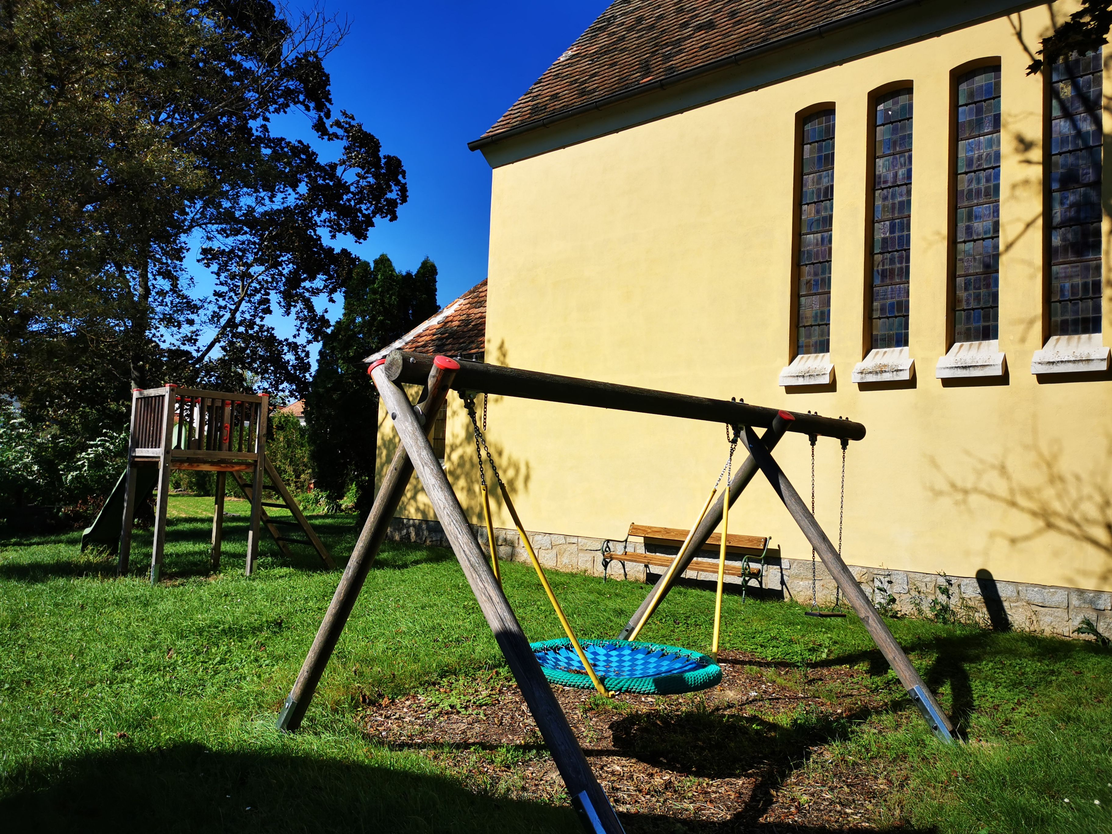 Spielplatz mit Schaukel und Rutsche neben einer gelben Kapelle mit hohen Fenstern.