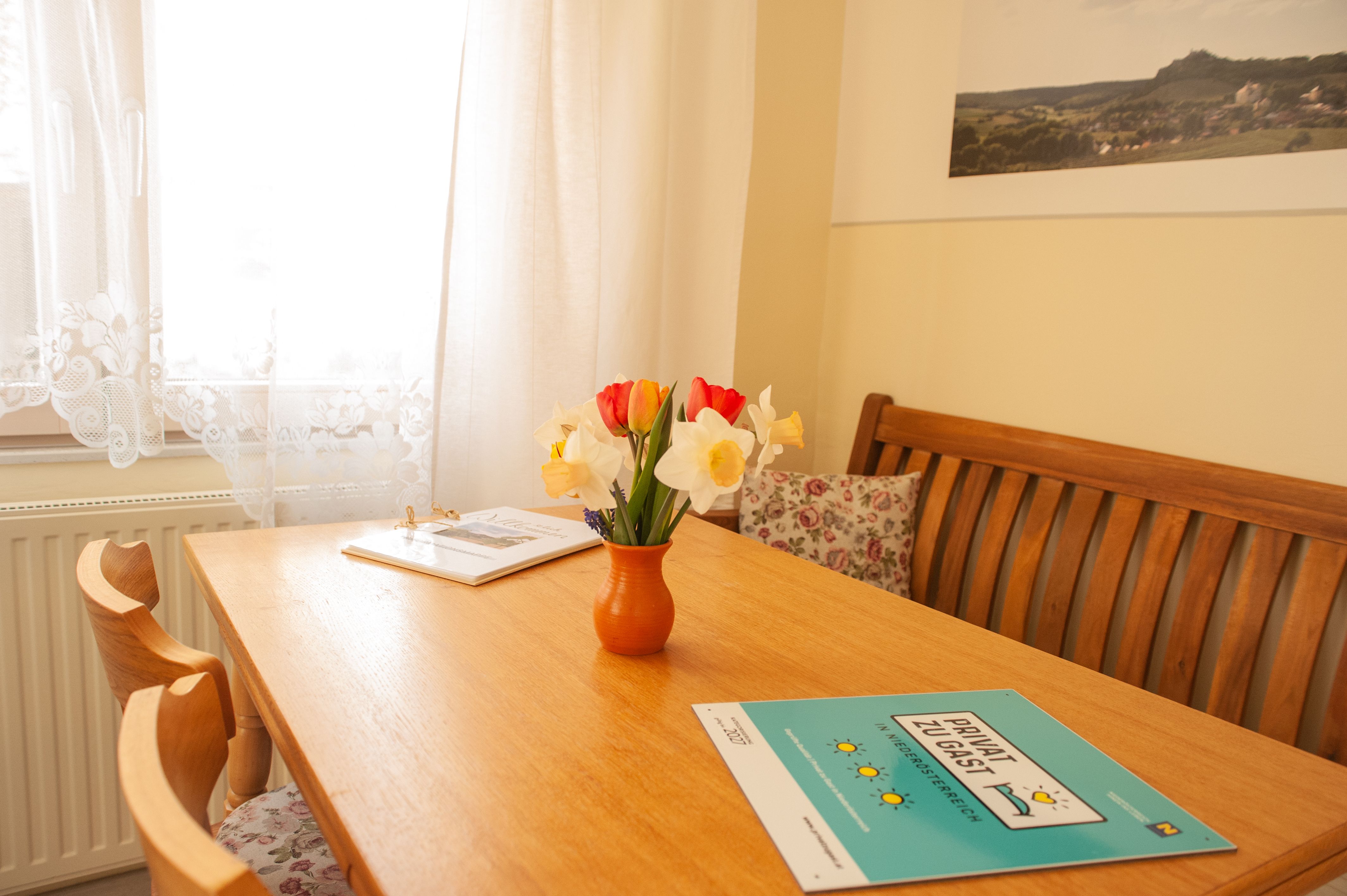 Útulná jedáleň s dreveným stolom, kyticou kvetov vo váze a brožúrami. Svetlé závesy a obraz na stene.