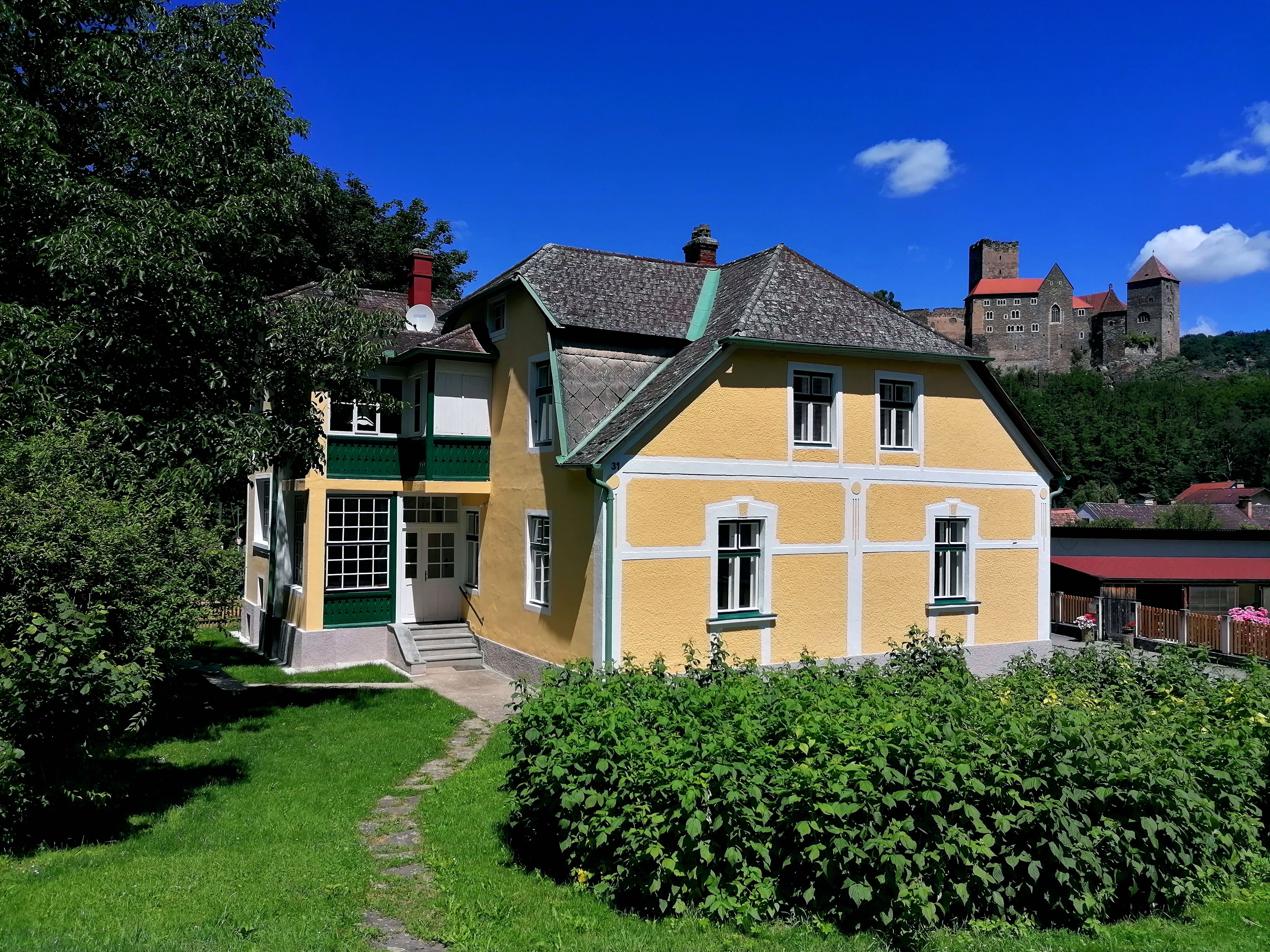 Gelbes Haus mit grünem Garten, im Hintergrund eine Burg auf einem Hügel.