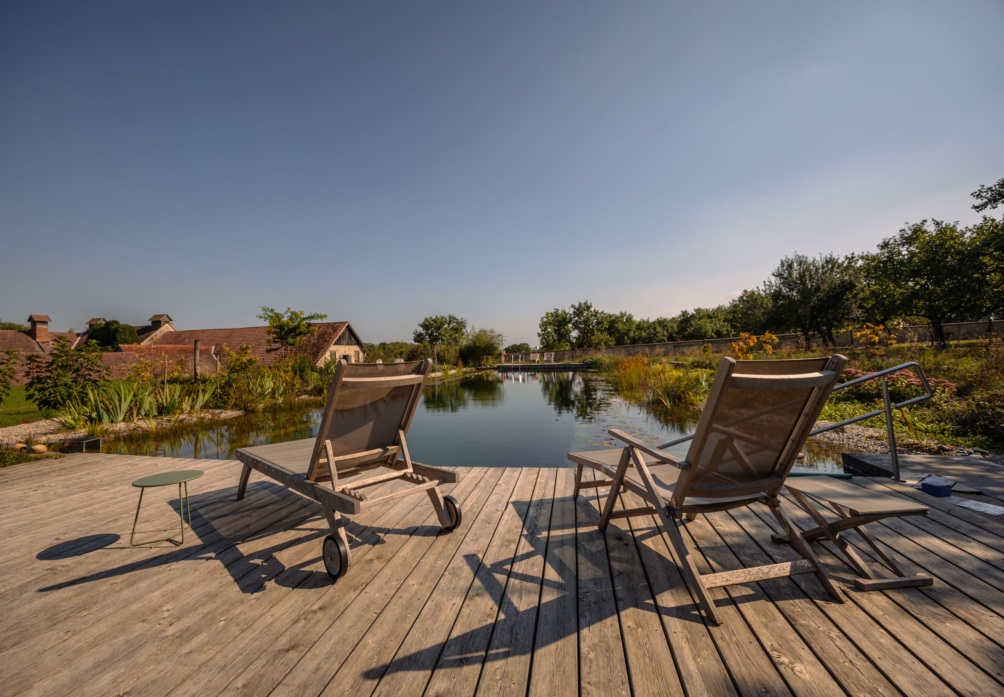 Zwei Liegestühle auf einem Holzdeck am Teich mit Garten im Hintergrund.