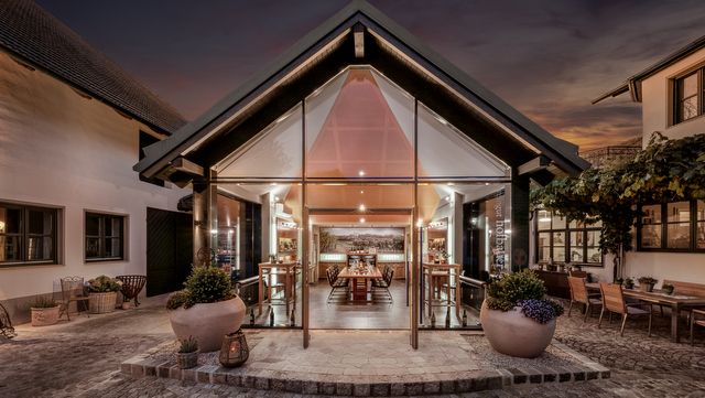 Moderner Verkostungsraum mit Glasfront und Holztischen, umgeben von traditionellen Gebäuden bei Dämmerung.