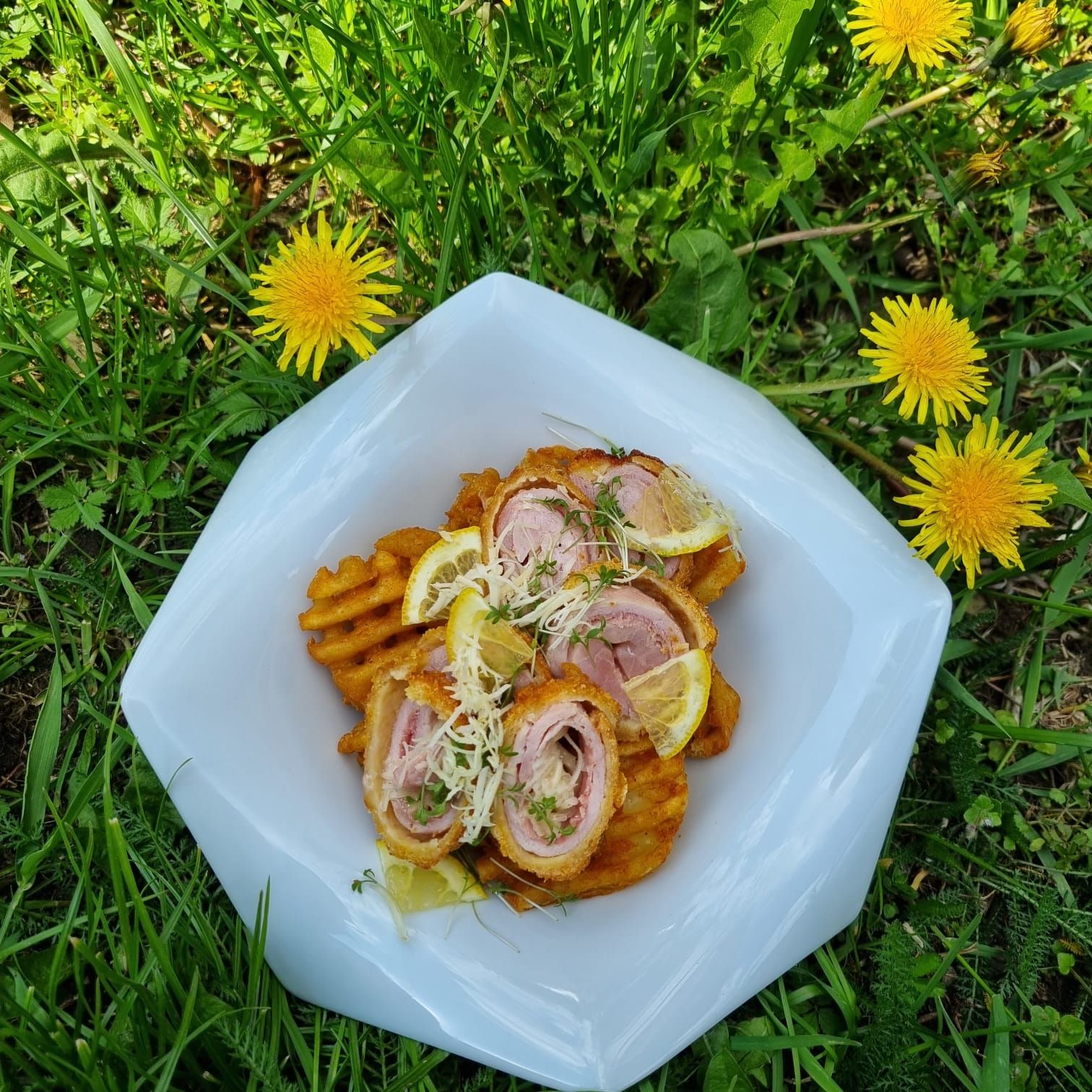 Ein Teller mit Cordon Rolls und Waffelpommes, garniert mit Zitronenscheiben und Kresse, liegt auf einer Wiese mit Löwenzahn.