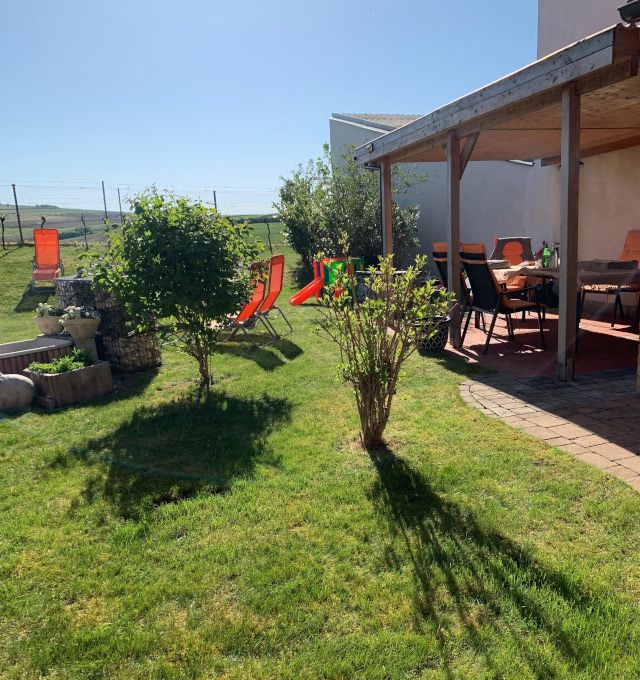 Ein sonniger Garten mit Rasen, Sträuchern, Liegestühlen und einer überdachten Terrasse mit Tisch und Stühlen.