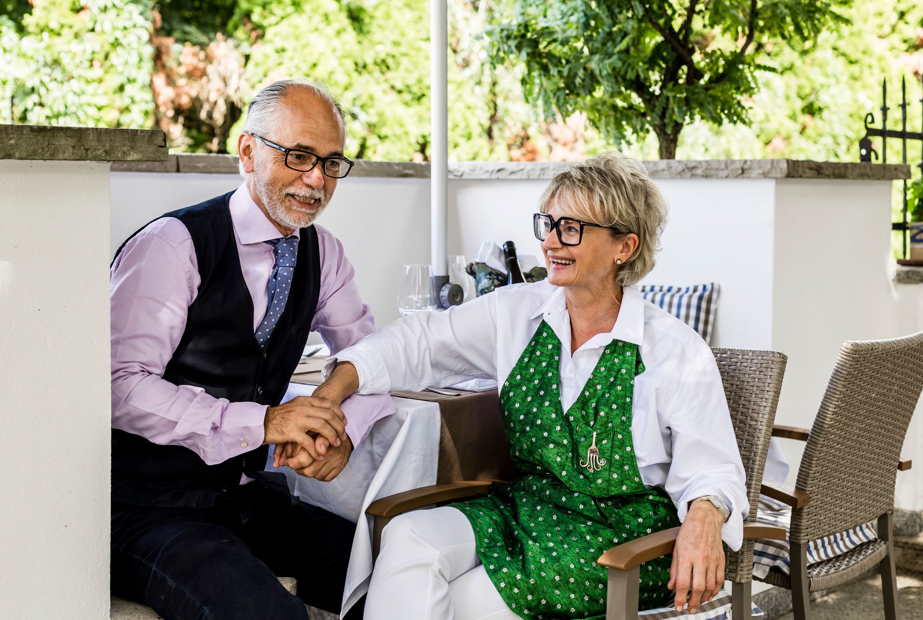 Ein Mann und eine Frau sitzen lächelnd an einem Tisch im Freien, sie halten sich an den Händen.