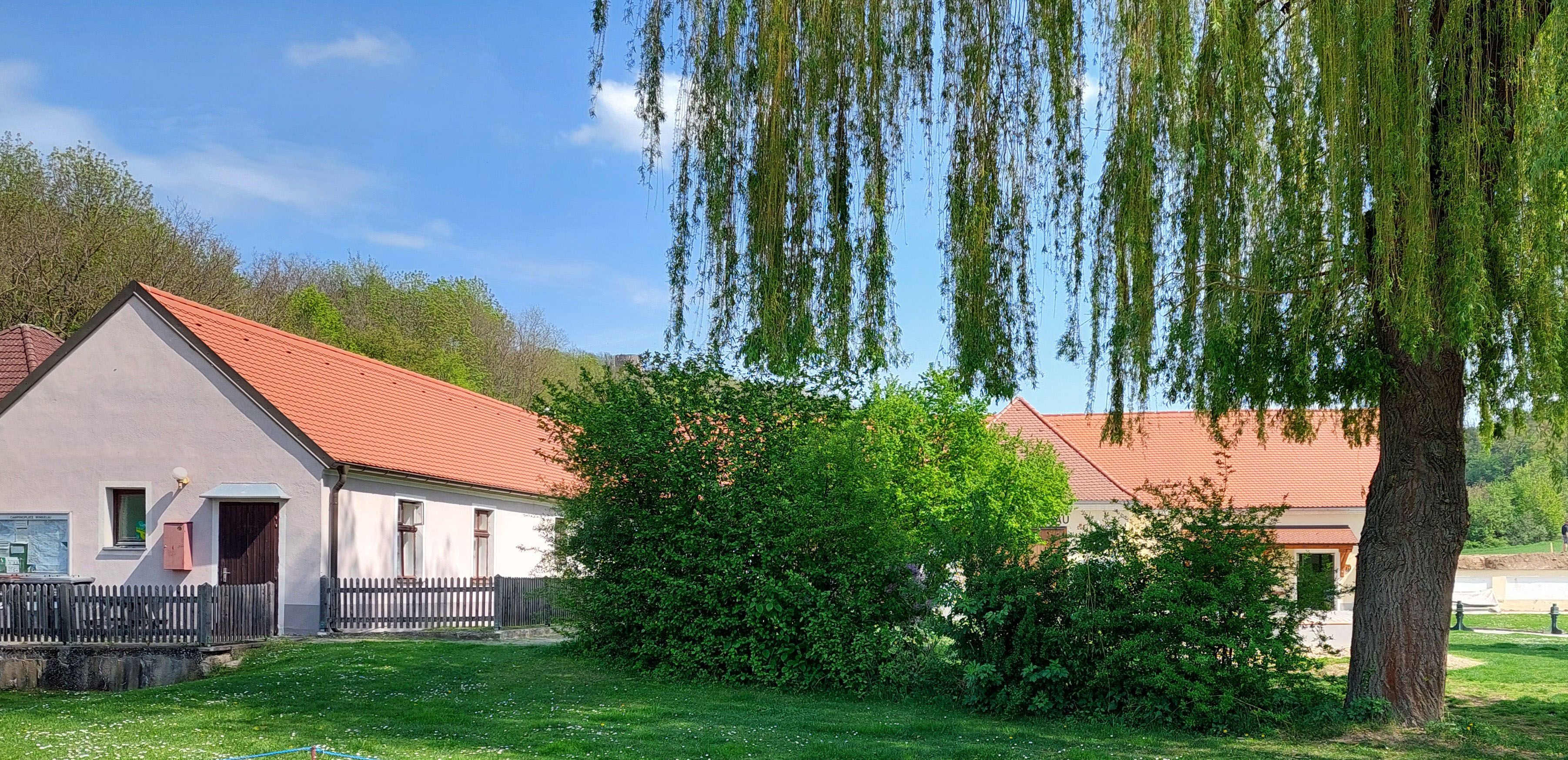 Ein ländliches Gebäude mit roten Ziegeldächern, umgeben von Bäumen und Rasen, unter einem blauen Himmel.