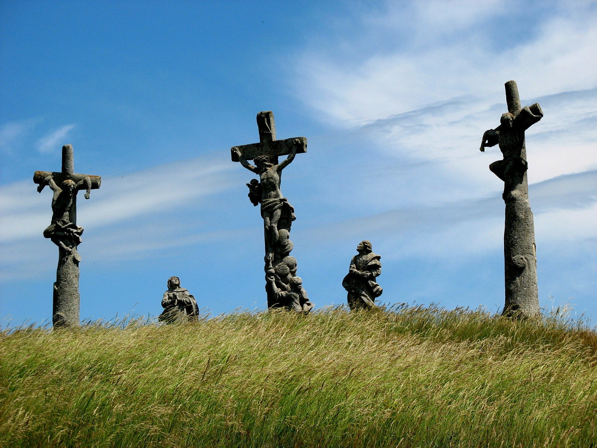 Drei Kreuze mit Figuren auf einem Hügel unter blauem Himmel.