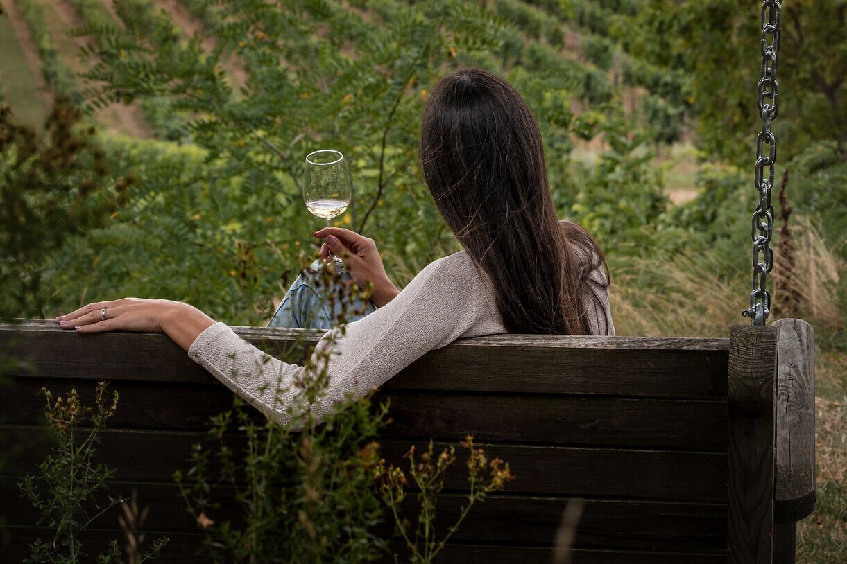 Eine Frau sitzt auf einer großen Holzschaukel und genießt den Ausblick auf die Weinviertler Landschaft mit zahlreichen Weingärten. In der Hand hält sie ein Glas Weißwein.