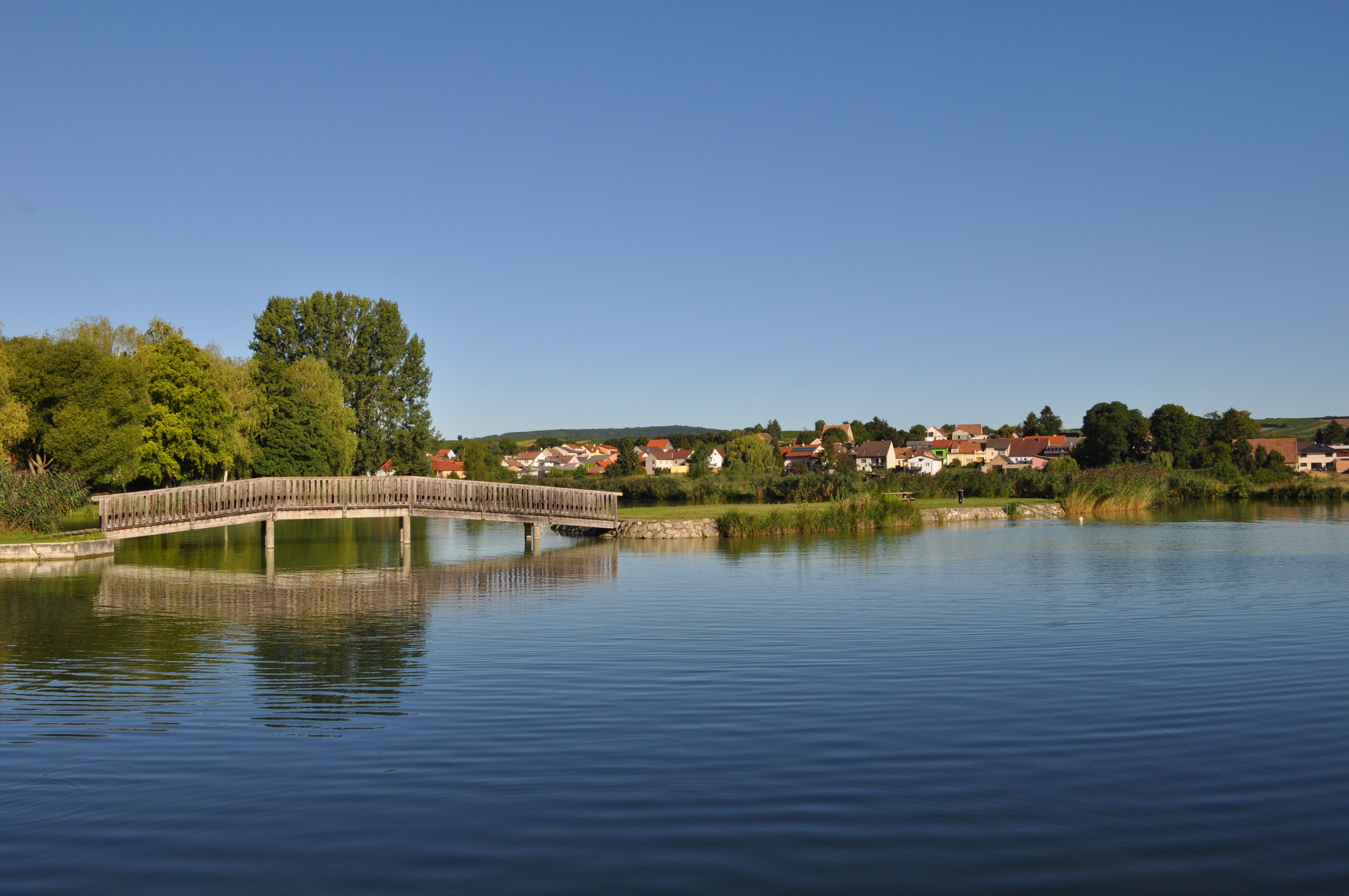 Holzbrücke über einen Teich mit Dorf im Hintergrund.