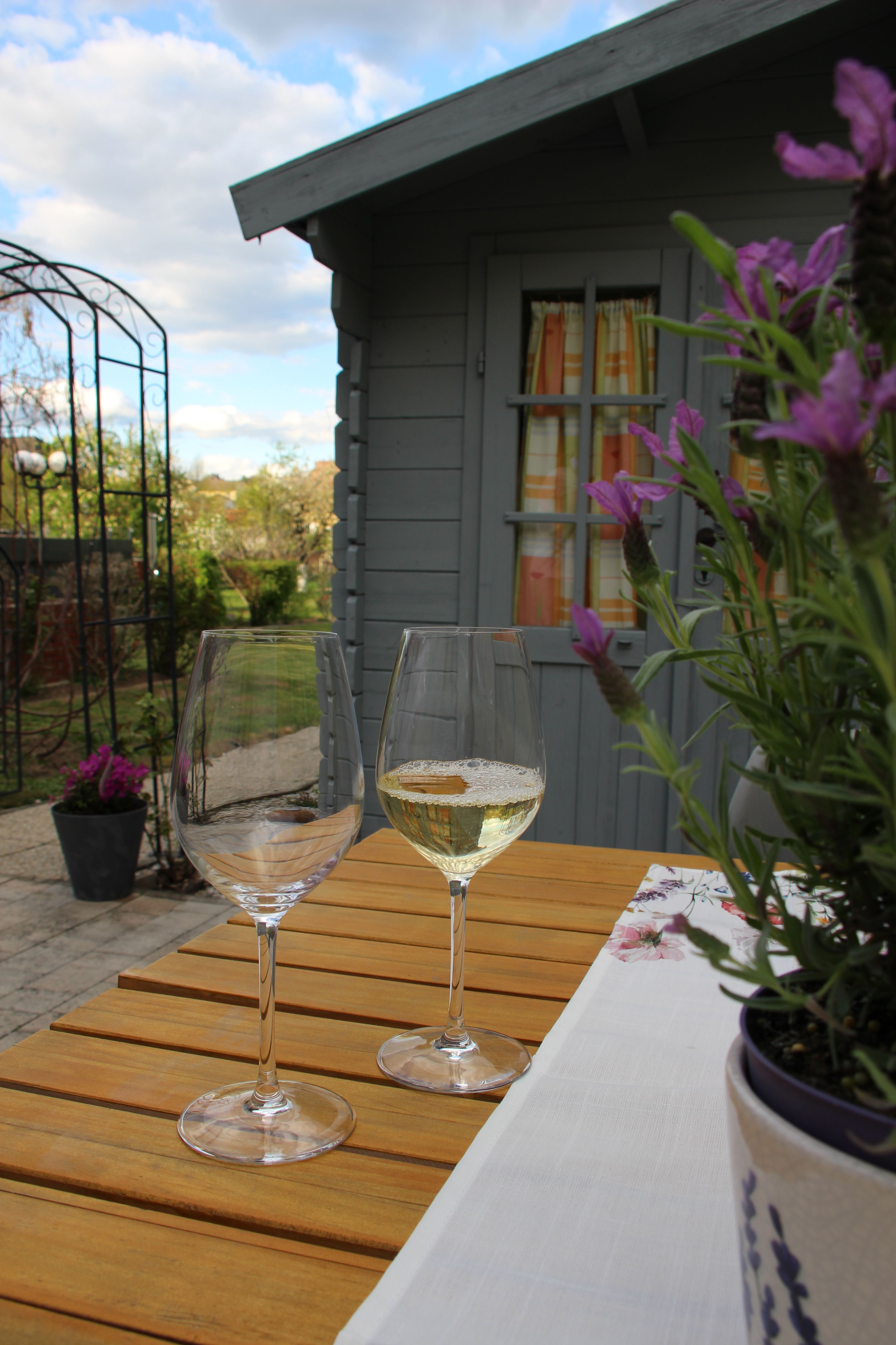 Two wine glasses on a wooden table in front of a garden house with flowers in the foreground.
