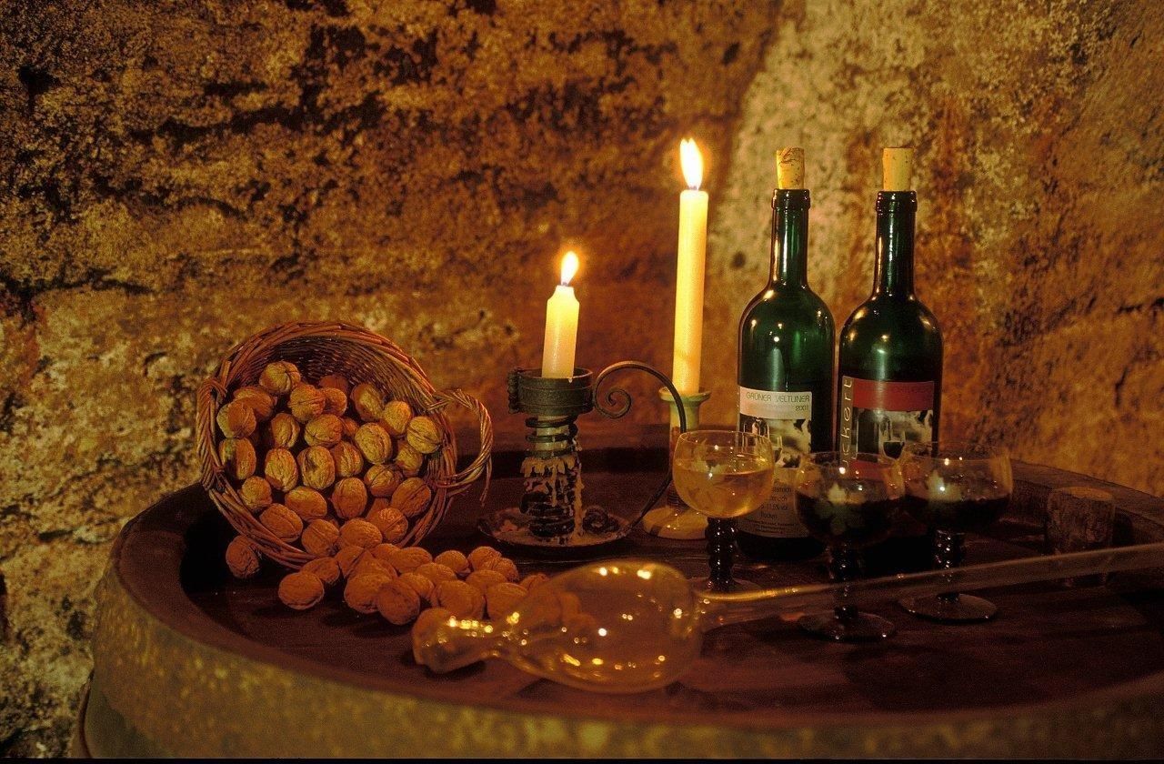 Ein Weinkeller mit Kerzenlicht, zwei Weinflaschen, Gläsern und einem Korb voller Walnüsse auf einem Fass.