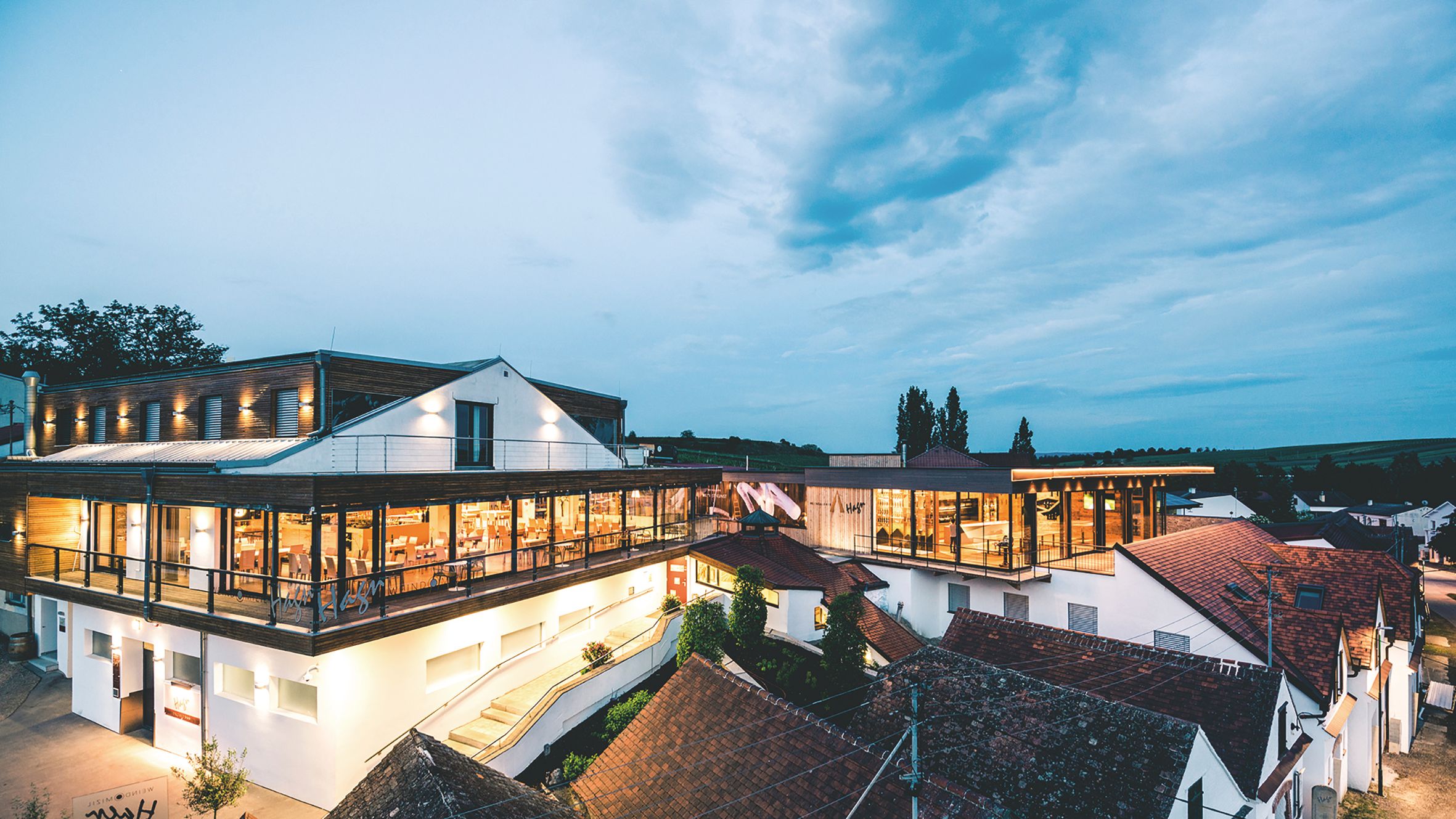 Moderne Weingut-Architektur bei Dämmerung mit beleuchteten Innenräumen und umliegenden Gebäuden.