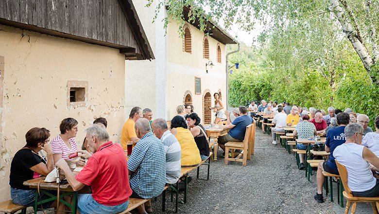 Lidé sedí u dlouhých stolů venku v heurigeru.