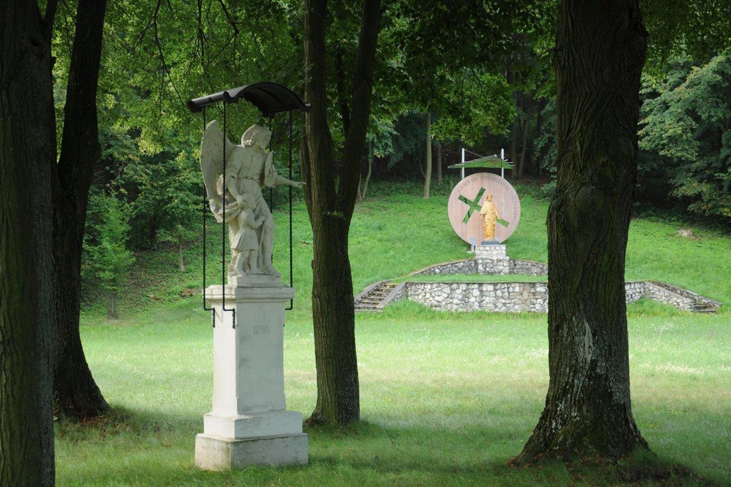 Statue eines Engels mit Kind im Vordergrund, im Hintergrund eine weitere Statue auf einem Podest in einem bewaldeten Park.