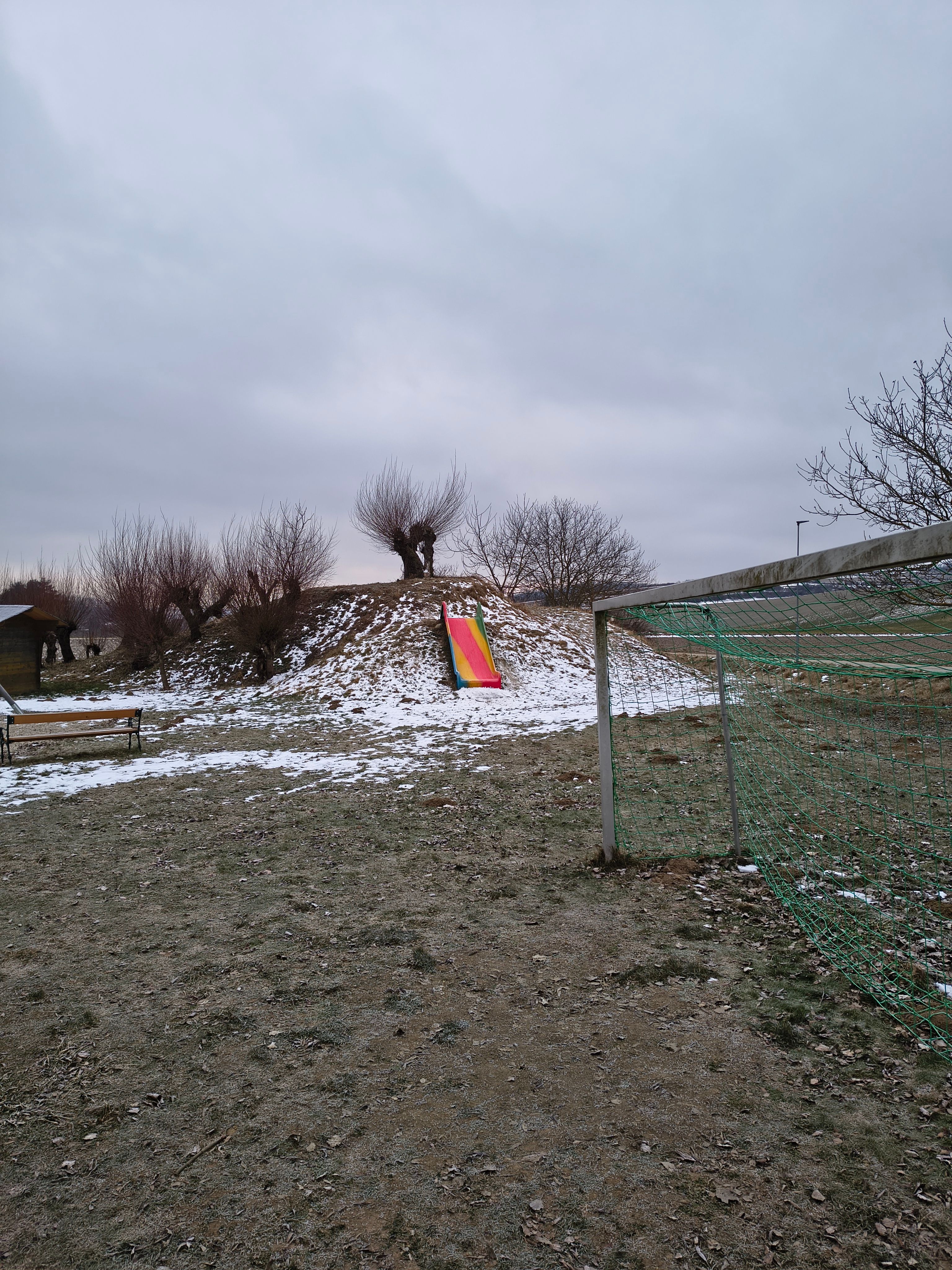 A playground with a colorful slide on a hill, surrounded by bare trees and a soccer goal in the foreground.