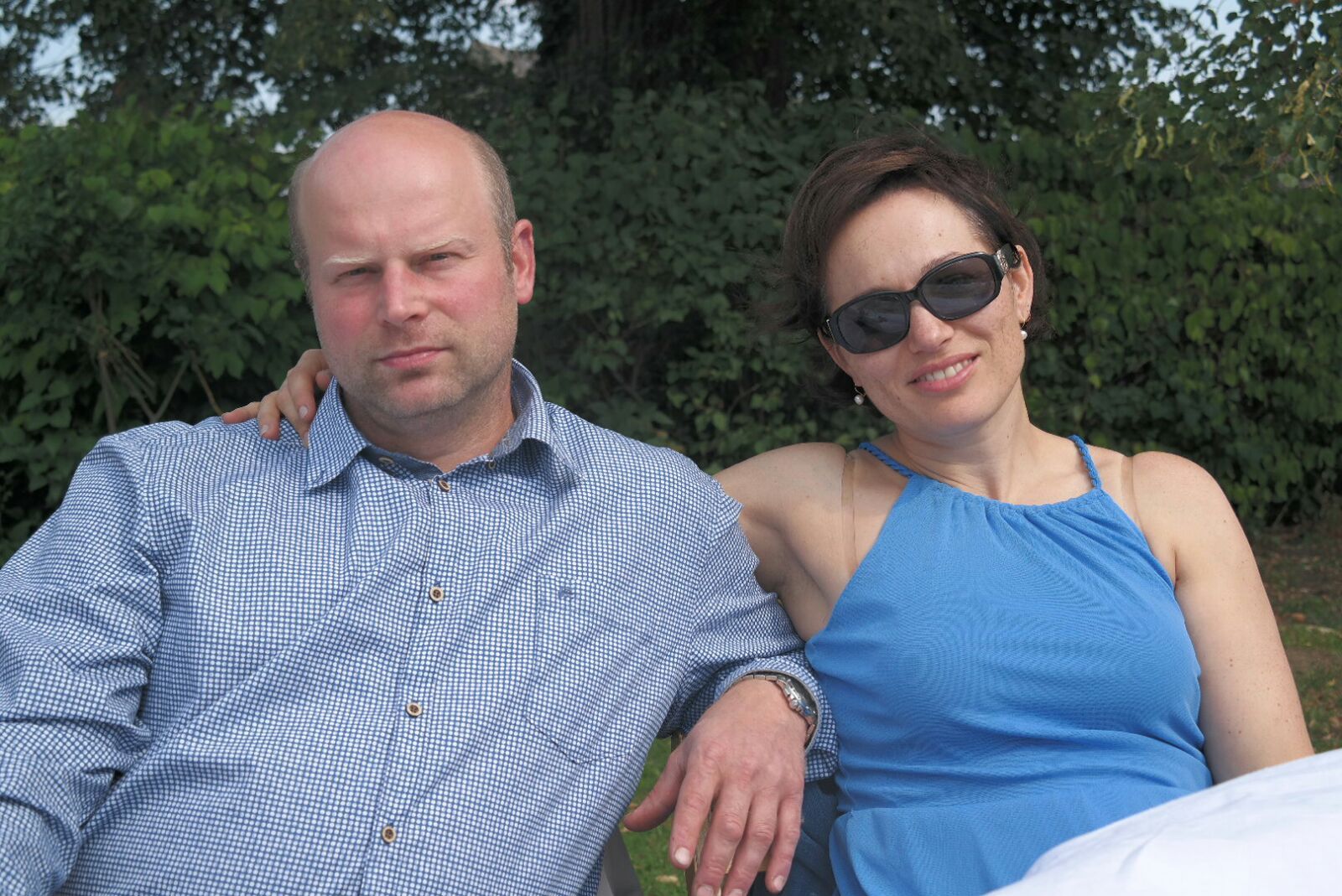 A man and a woman are sitting next to each other outdoors, the woman is wearing sunglasses.