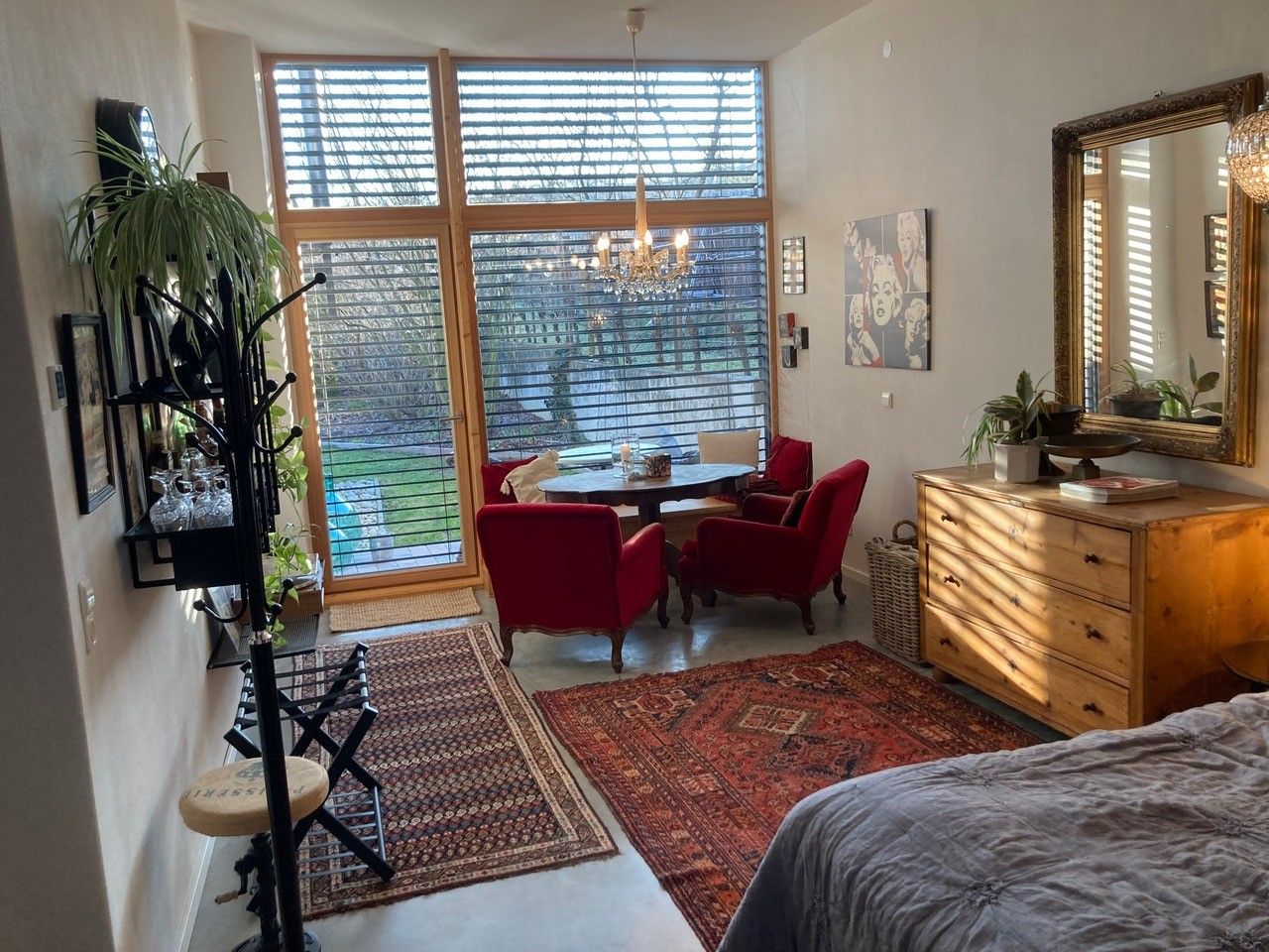 Wohnzimmer mit großen Fenstern, roten Sesseln, Holzmöbeln und Teppichen, Blick in den Garten.