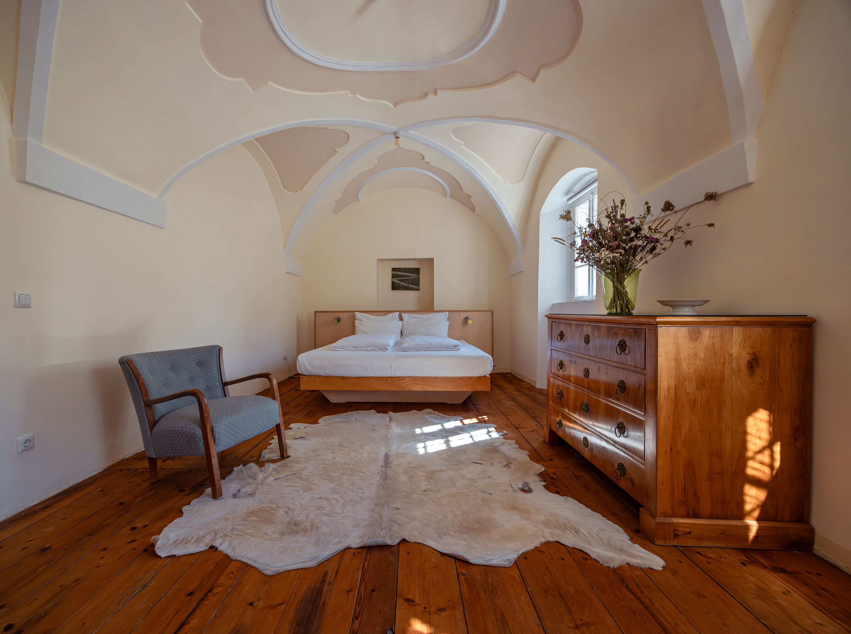 Schlafzimmer mit Gewölbedecke, Holzboden, Bett, Sessel, Kommode und Fellteppich.