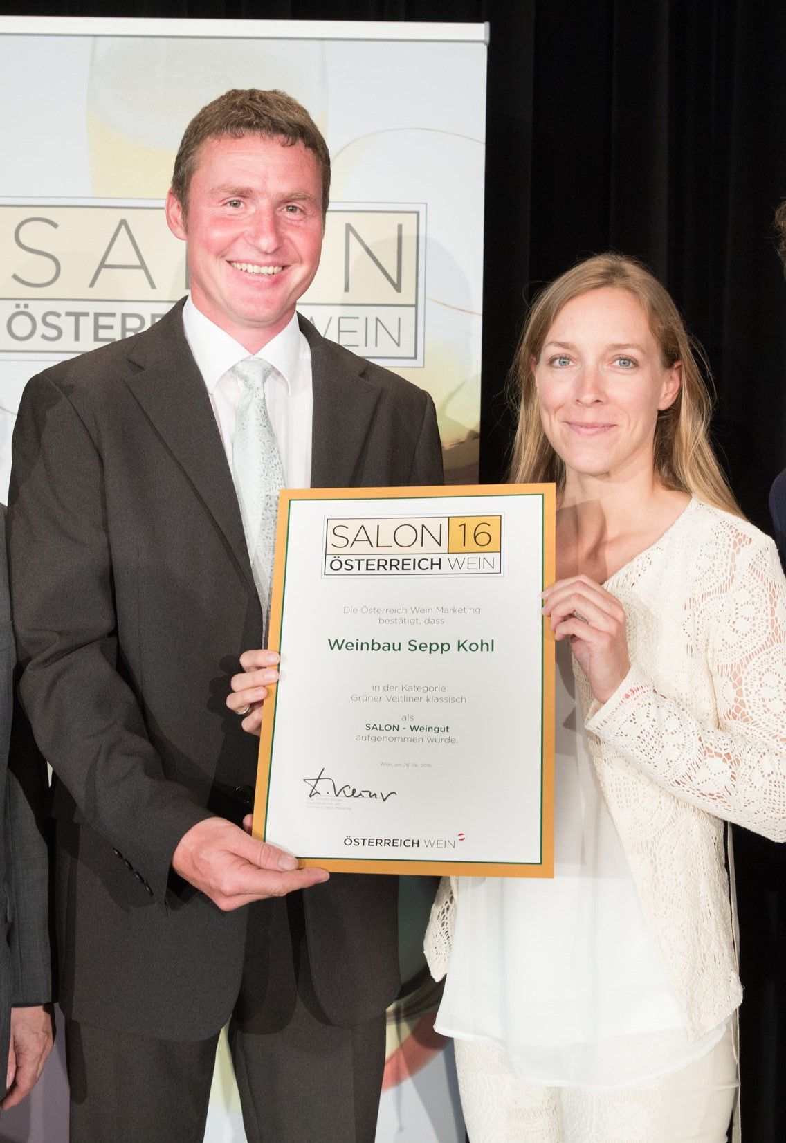 Zwei Personen halten eine Urkunde mit der Aufschrift 'Weinbau Sepp Kohl' bei einer Veranstaltung.