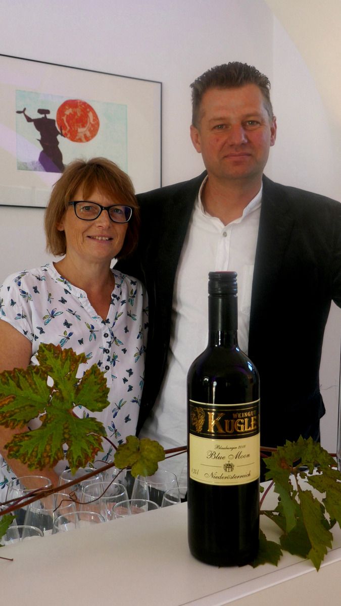 A man and a woman stand behind a bottle of wine from the Kugler winery. Vine leaves can be seen in the foreground.