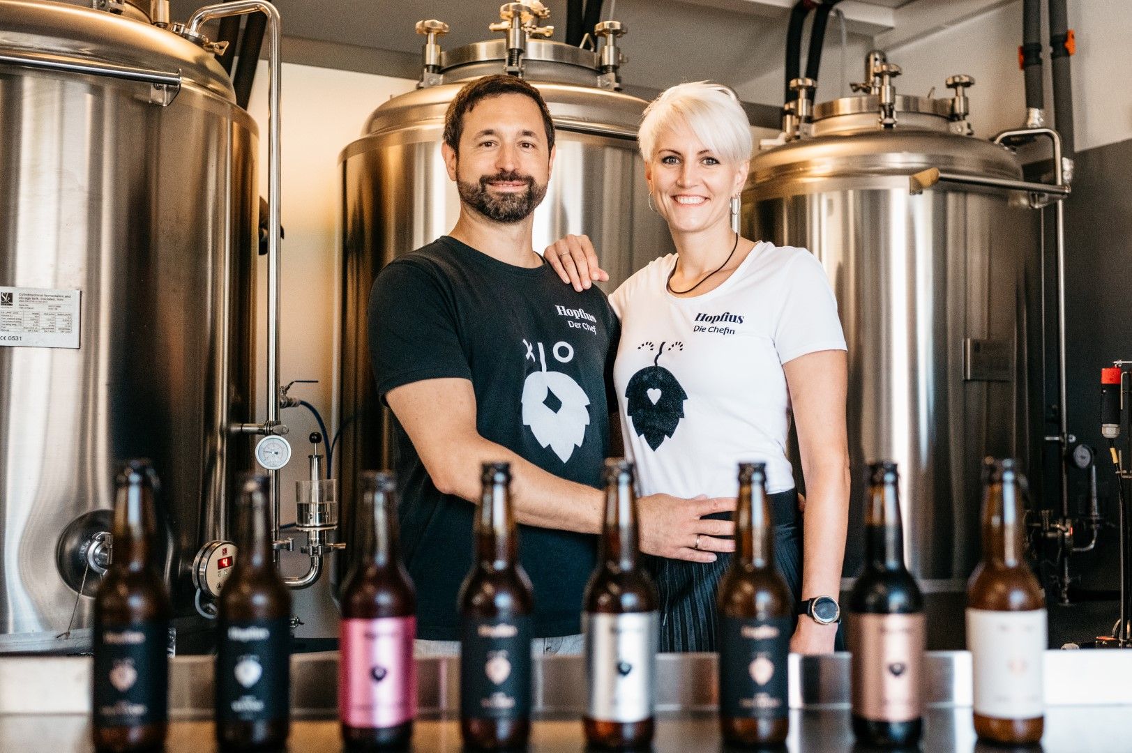 Ein Mann und eine Frau stehen lächelnd vor Brauereitanks, mit verschiedenen Bierflaschen im Vordergrund.