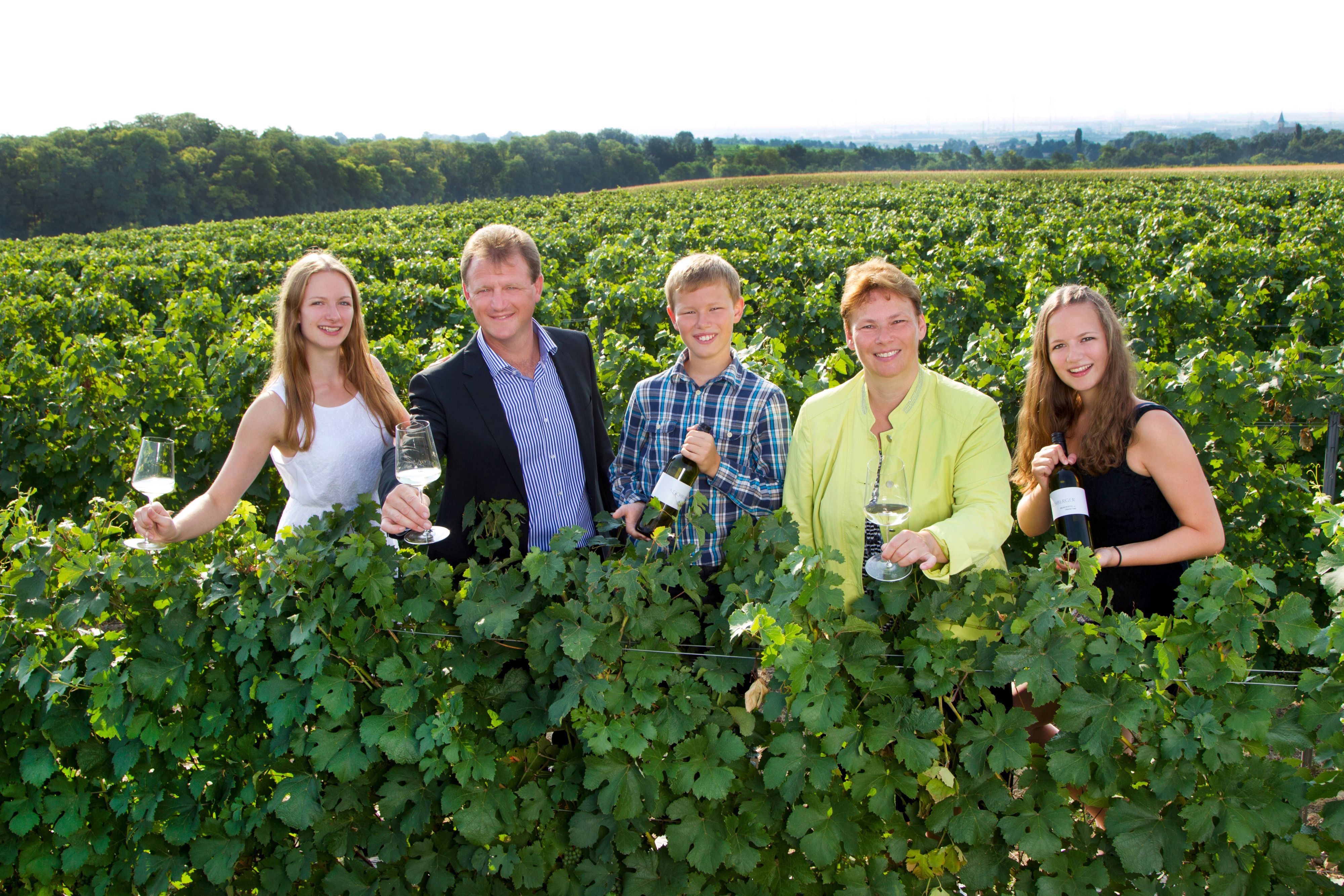 A group of five people stand in a vineyard holding wine glasses and bottles.