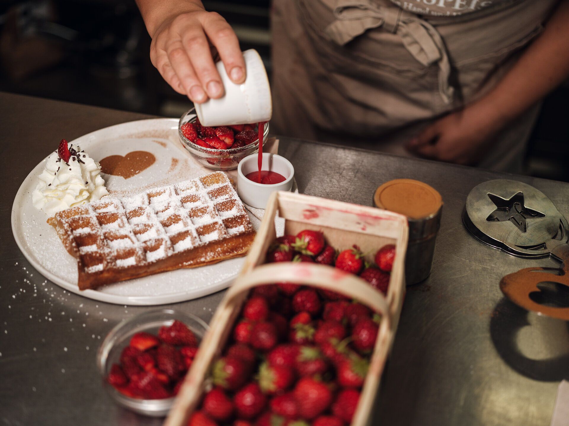 Ein Korb voller Erdbeeren und eine frische Waffel auf einem Teller, eine Hand gießt Erdbeersauce in ein kleines Gefäß.